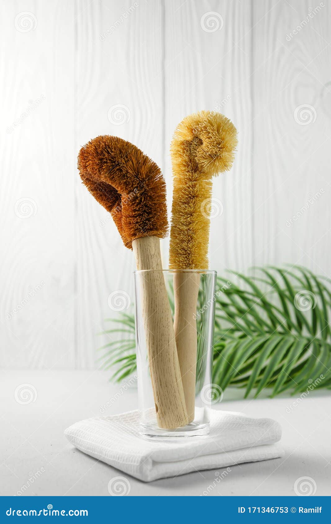 two scrapers made of coconut bristles and a bamboo handle in a glass cup on a light background.