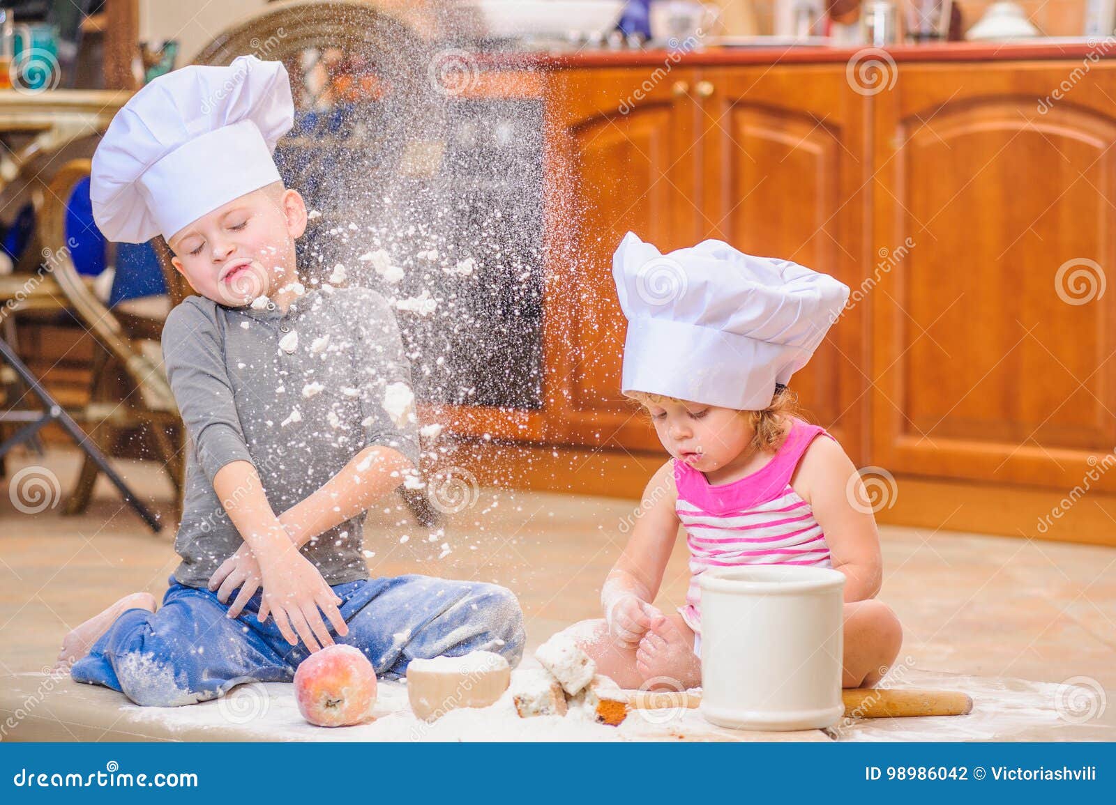 two siblings - boy and girl - in chef`s hats sitting on the kitchen floor soiled with flour, playing with food, making mess and ha