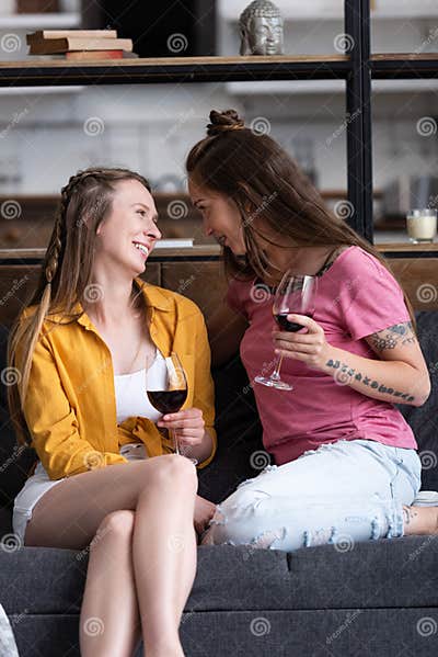 Two Smiling Lesbians Holding Wine Glasses And Looking At Each Other While Sitting On Sofa In