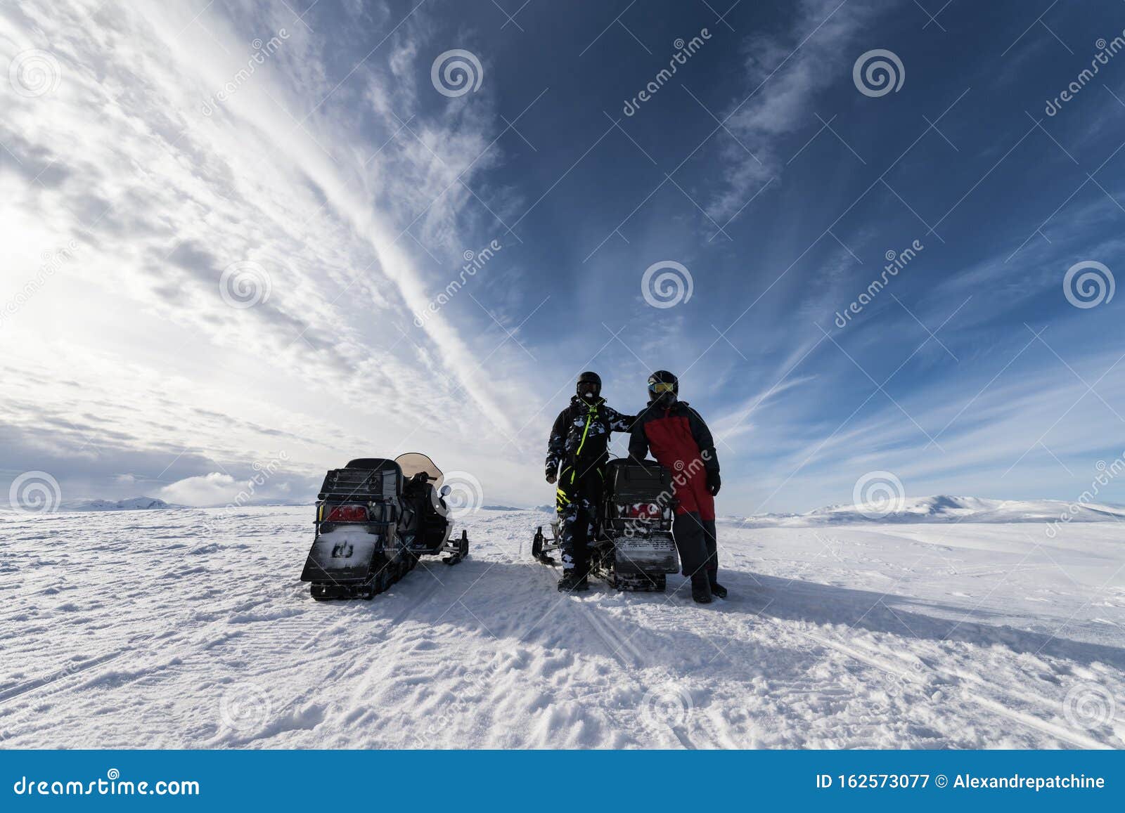 two snowmobiles back side and two friends pose around of one snowmobile. they dressed for driving in cold mountains