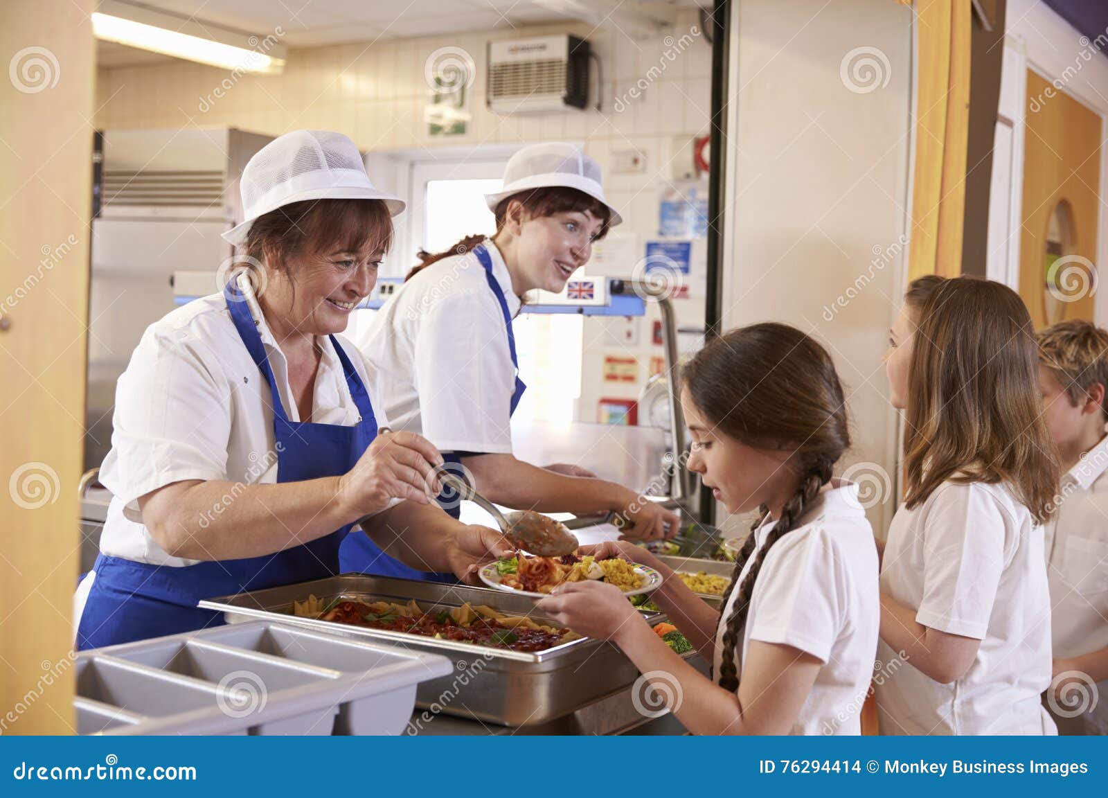 two women serving food to a girl in a school cafeteria queue