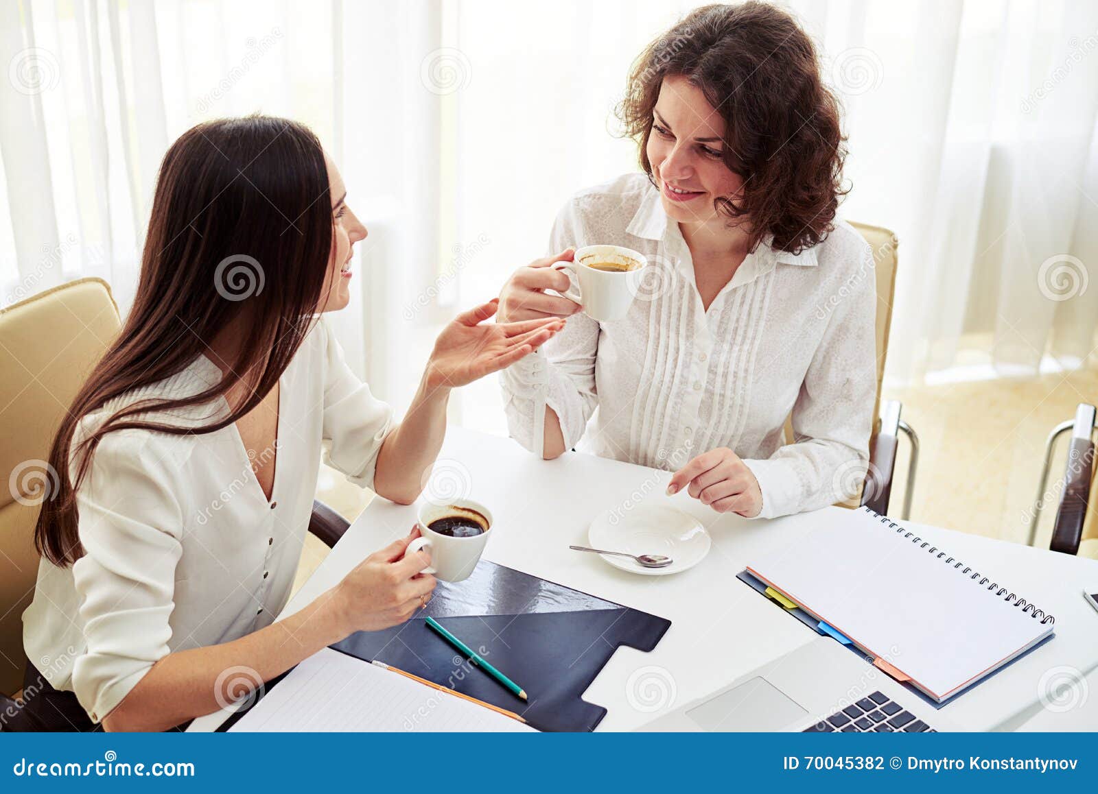 two women talking something drink coffee work young pretty hold hand spacious office 70045382