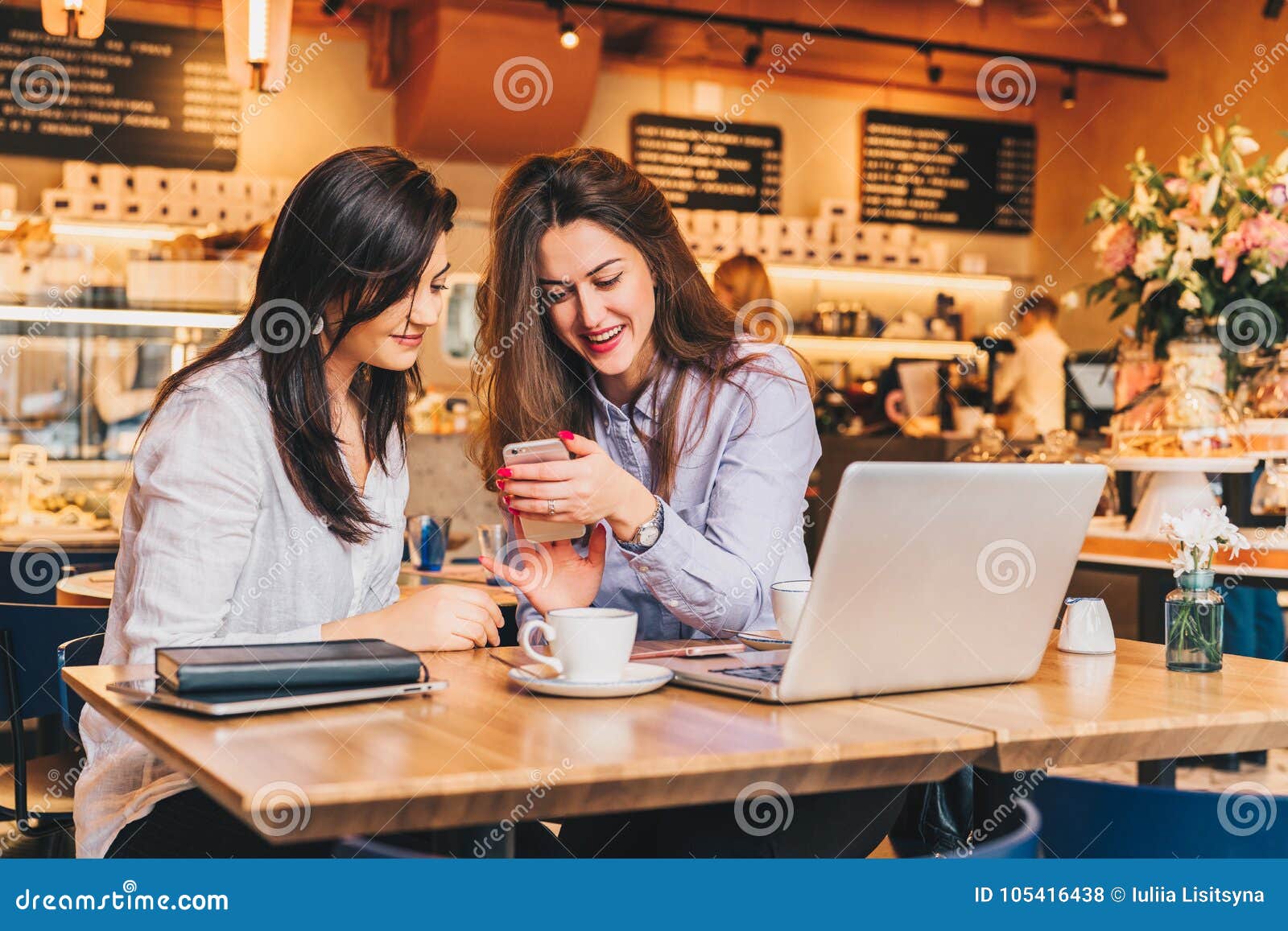 two young happy women are sitting in cafe at table in front of laptop, using smartphone and laughing.