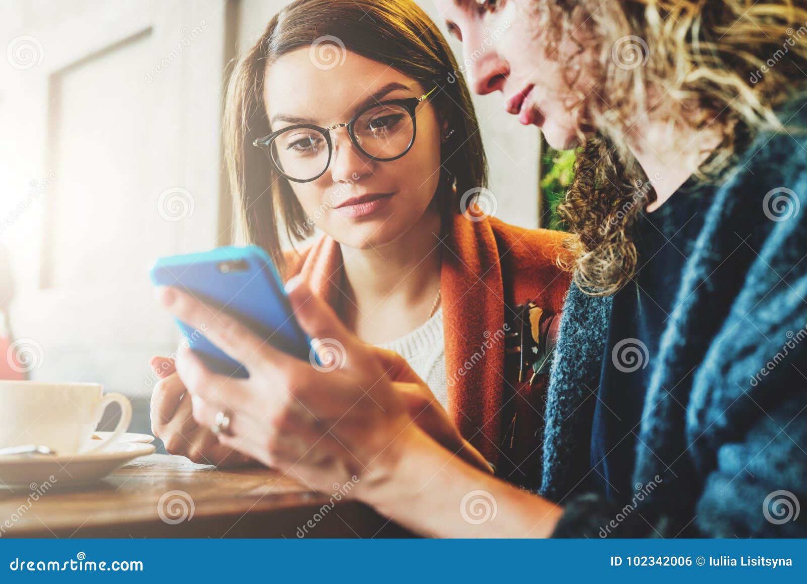 two young women sit in a cafe at the table and use a smartphone. the girl shows her friend a picture on the phone screen