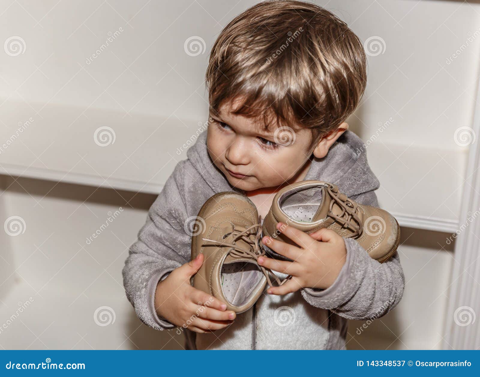 Un Niño Pequeño Con Una Cara Del Granuja Tiene Sus Pequeños Zapatos En Sus Manos Imagen de archivo - Imagen de divertido, zapatos: 143348537