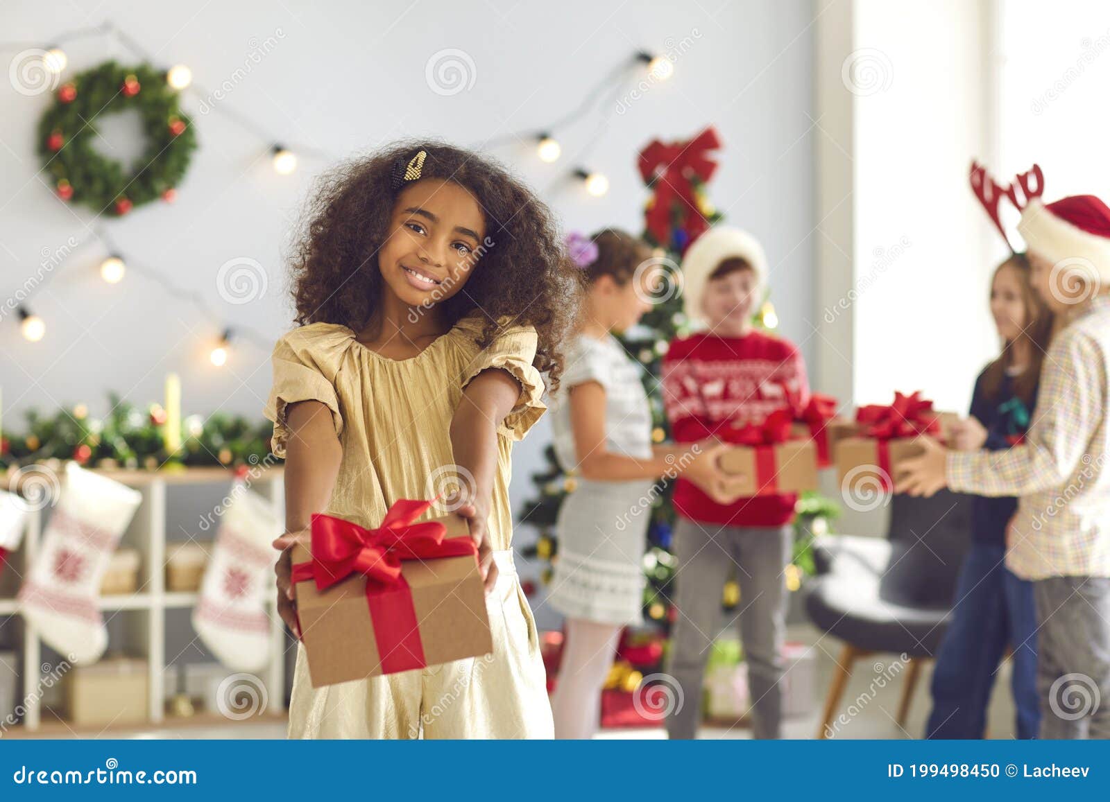 Una ragazza afro-americana che tiene per mano il regalo di Natale, guarda la macchina fotografica e sorride. Natale è condivisione Felice ragazza afro-americana che tiene per mano il presente e guarda la telecamera sullo sfondo sfuocato della stanza decorata con bambini che si scambiano regali