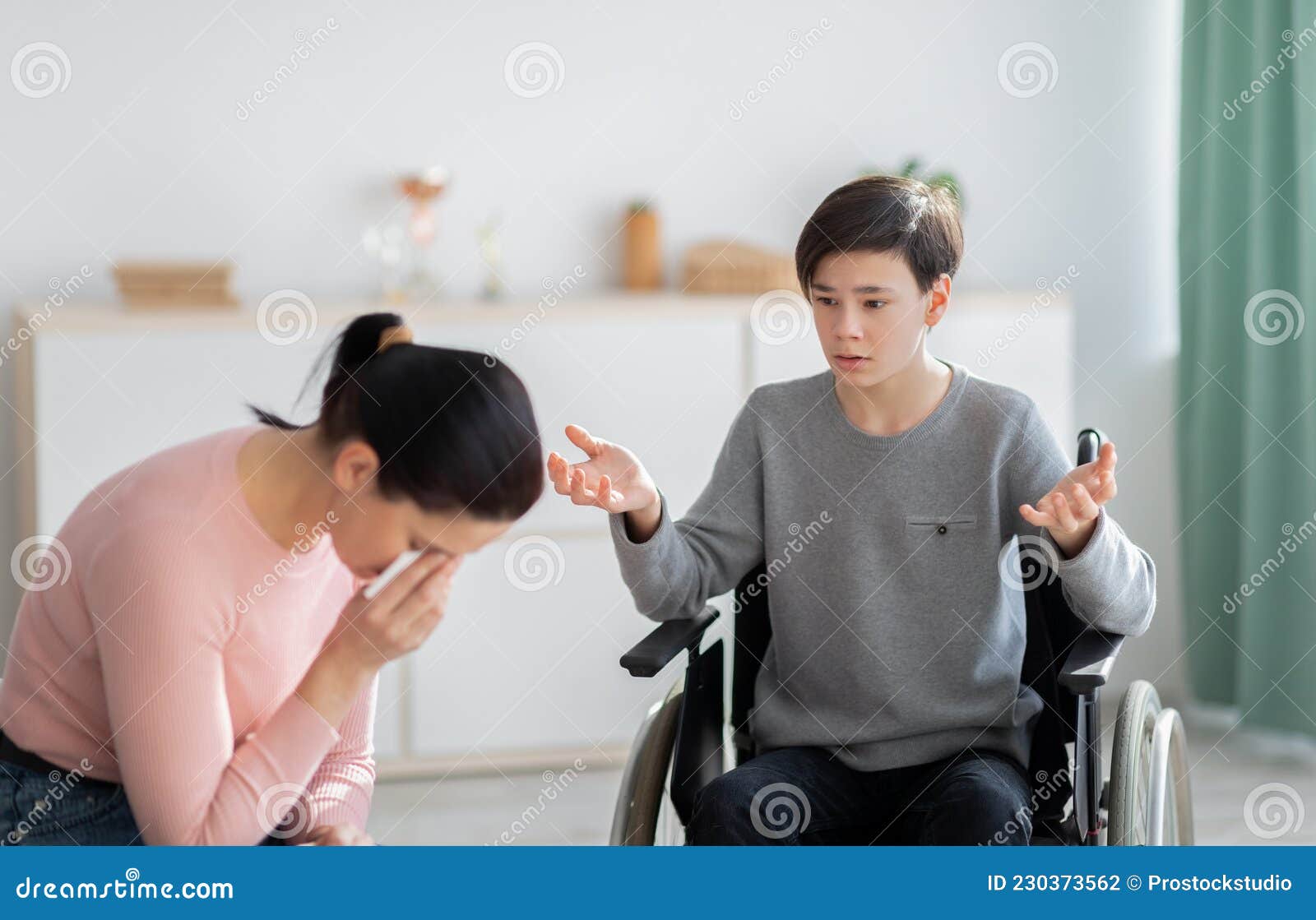 unhappy mother crying and her disabled teen son in wheelchair trying to justify himself after disagreement at home