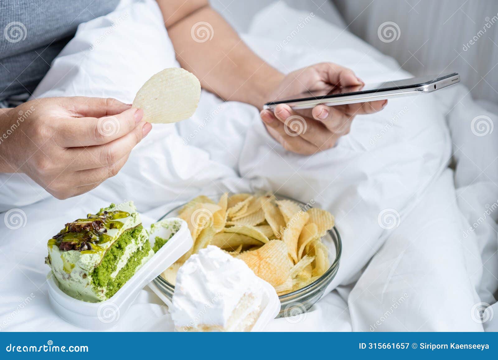 unhealthy woman eating junk food salty snack potato chips and dessert , cake in bed