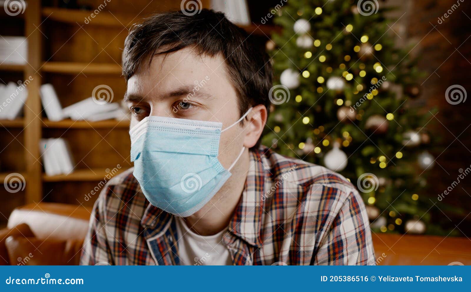 upset with bad mood man in protective mask is suffering from loneliness at home while sitting on couch near christmas tree during