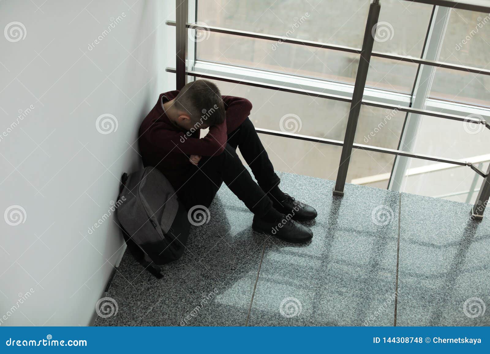 upset boy with backpack sitting near window indoors