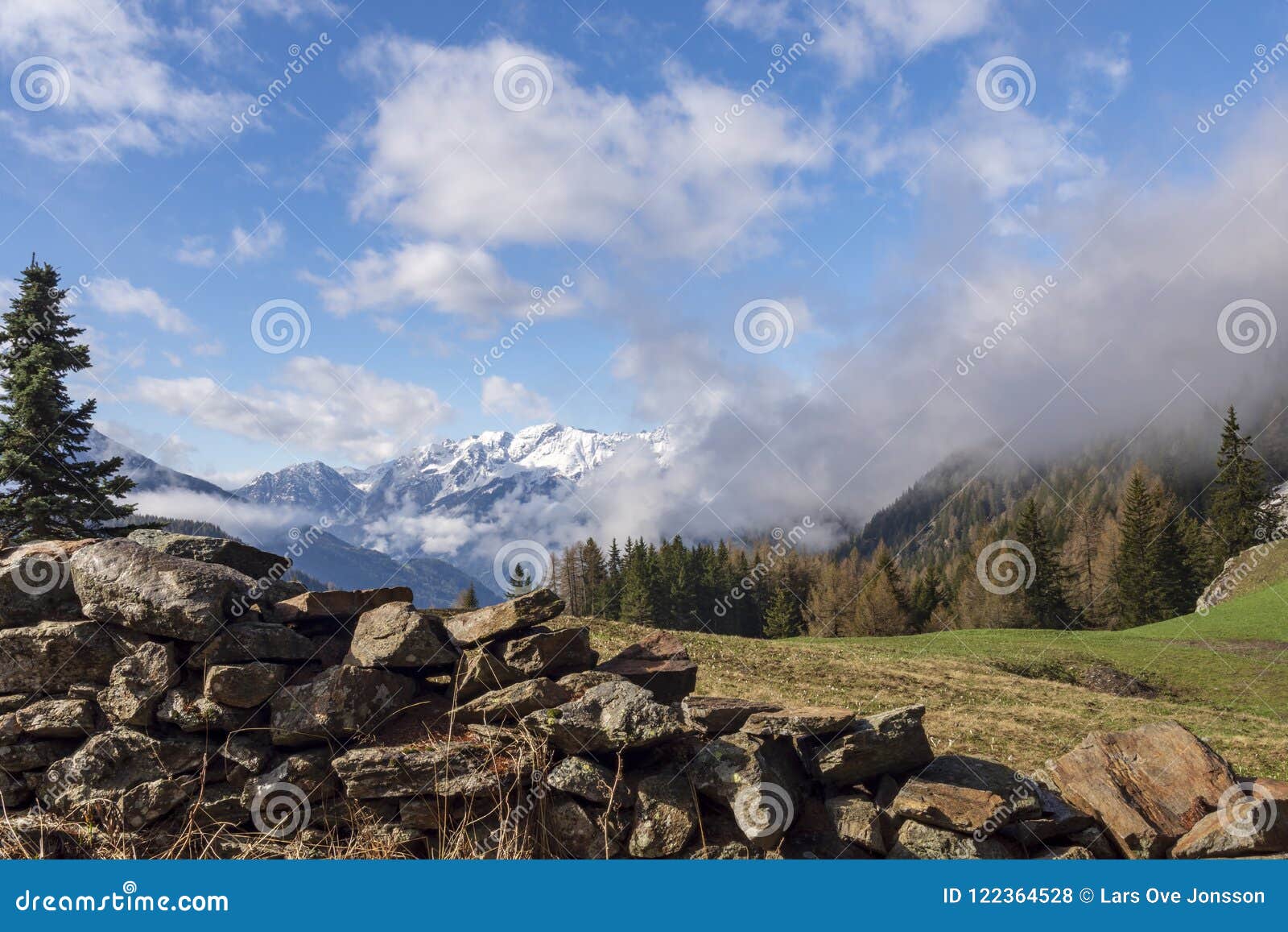 View Over Snowy Alps With An Old Stone Wall And A Green Meadow I Stock