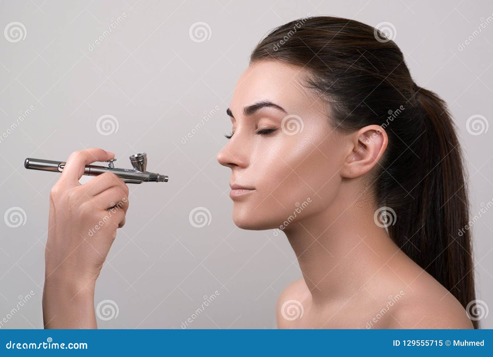 visagist making makeup for model with aerograph,  on grey. background with hand holding aerograph. visagist using aerograp