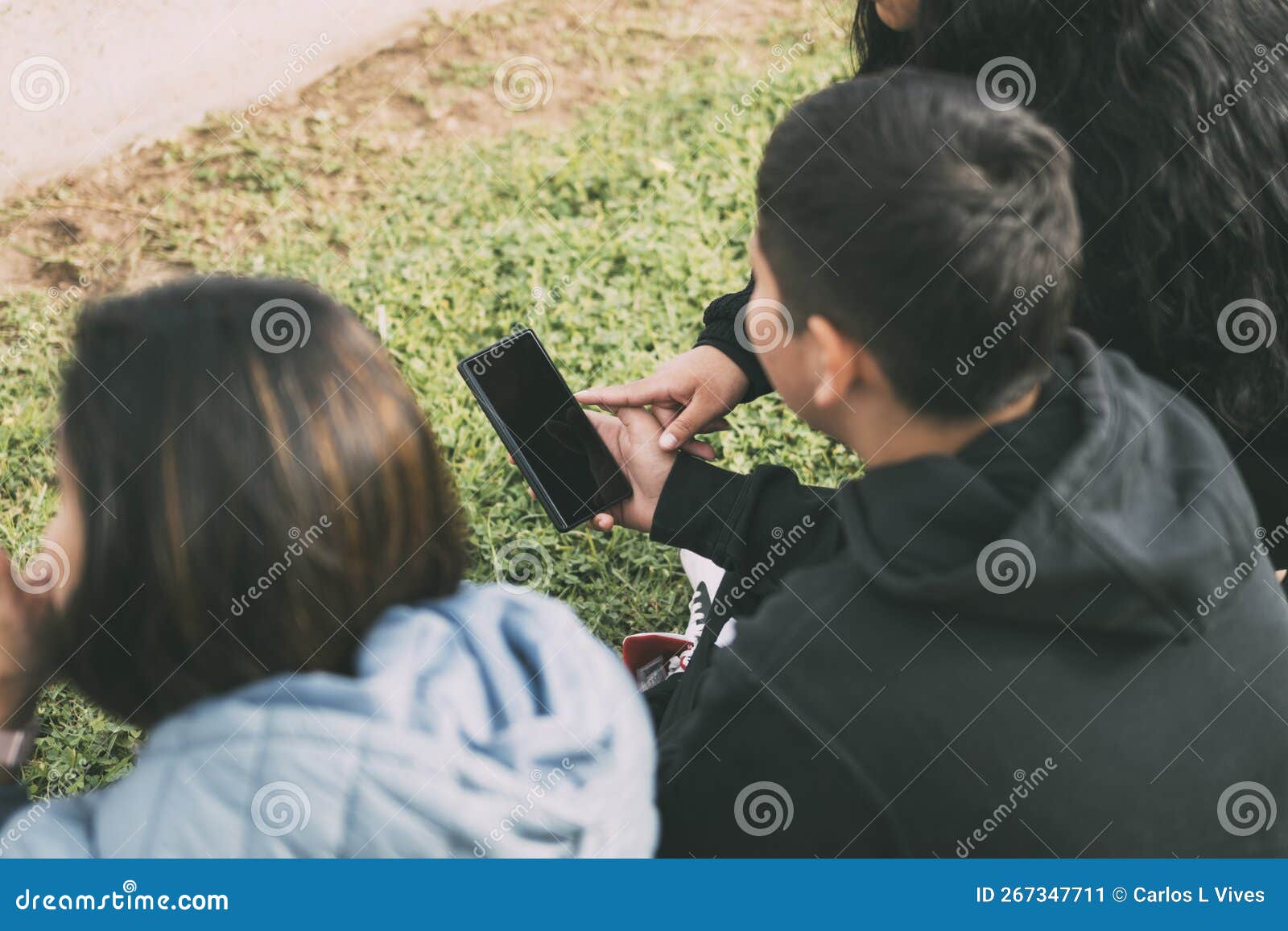 vista desde atras grupo de latinos riendo sentados en el suelo en un parque con un smartphone