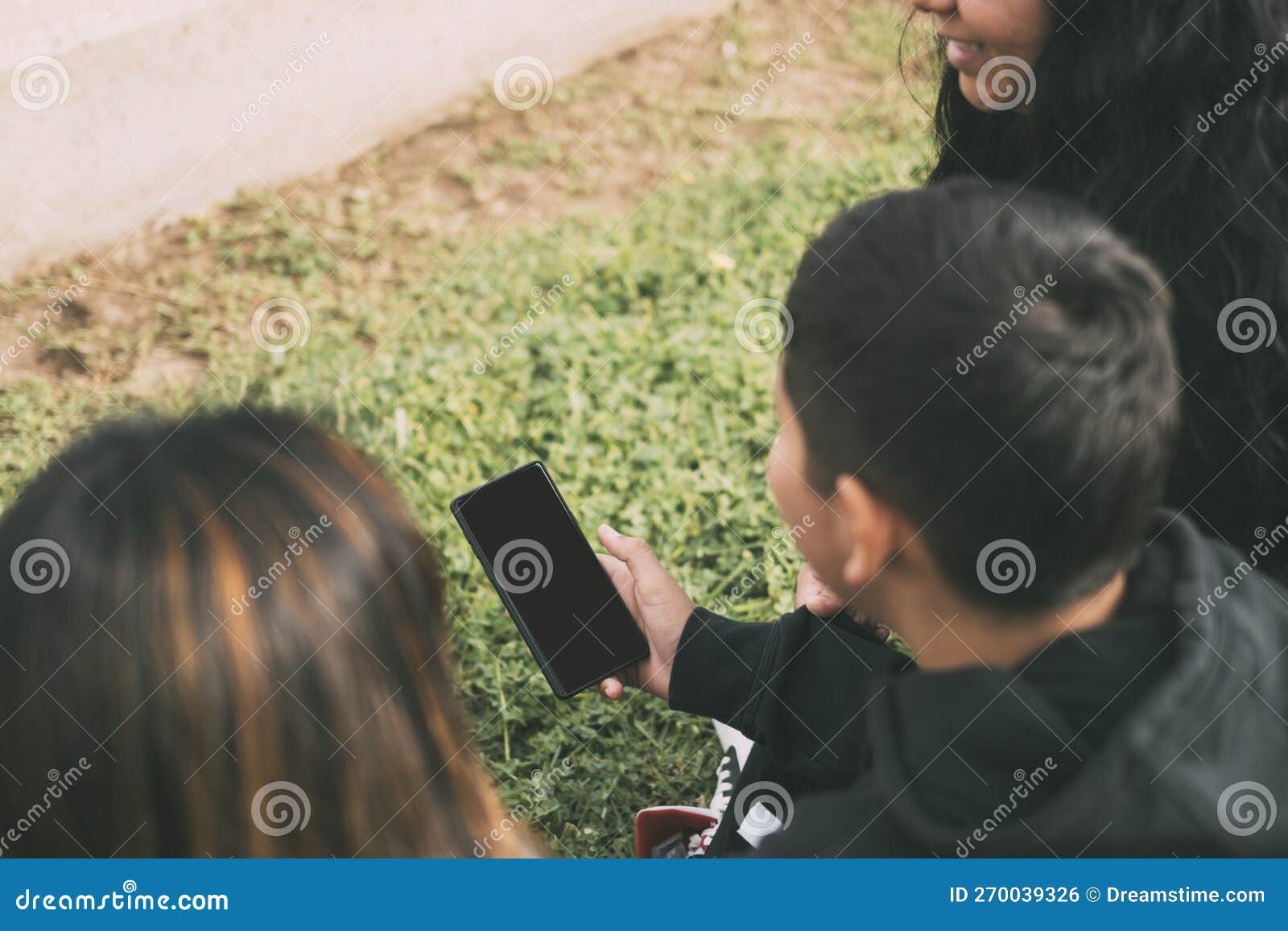 vista desde atras grupo de latinos riendo sentados en el suelo en un parque con un smartphone