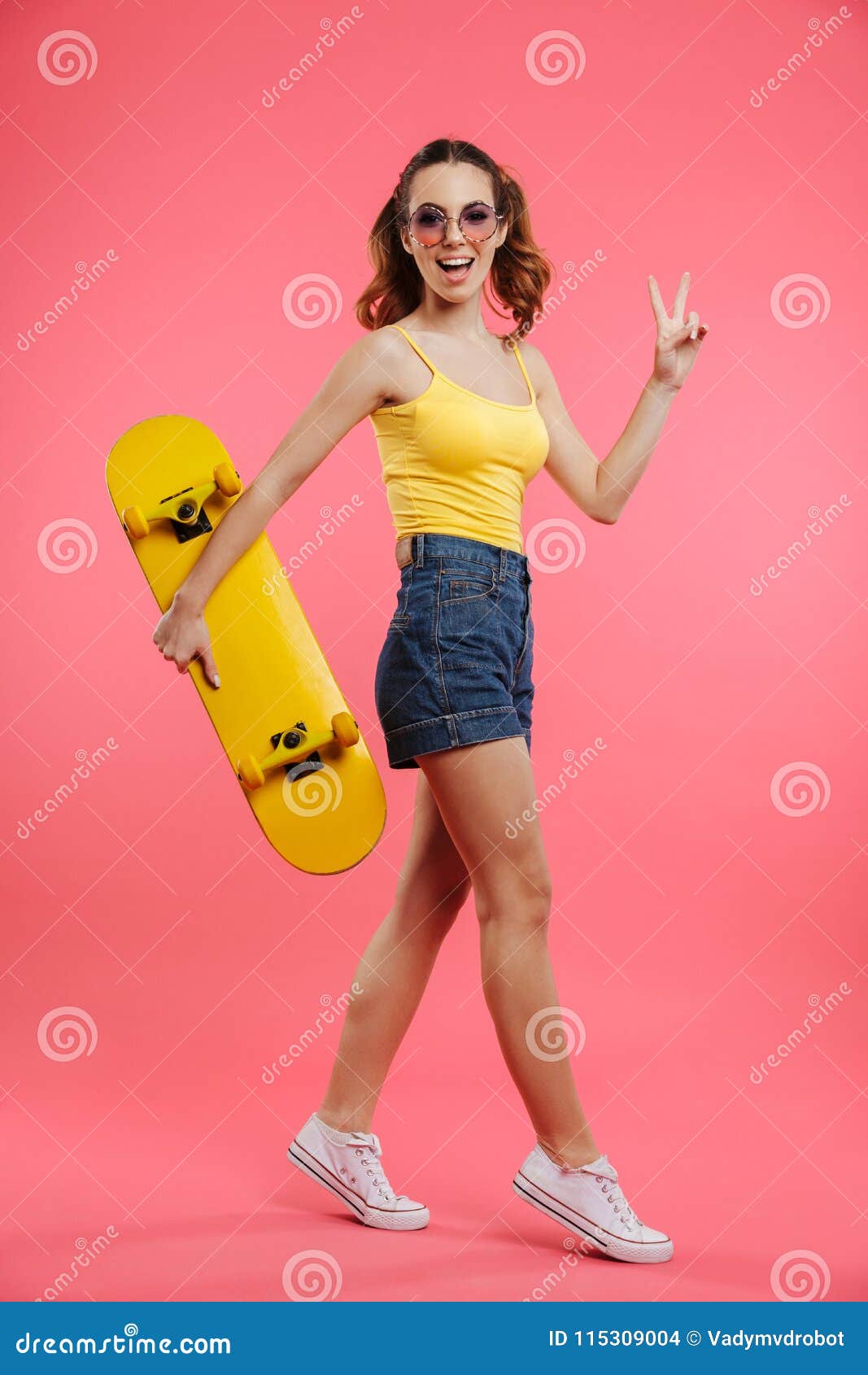 Volledig lengteportret van een vrolijk jong meisje. Het volledige lengteportret van een vrolijk jong meisje kleedde zich in de zomerkleren die skateboard houden en vredesgebaar over roze achtergrond tonen