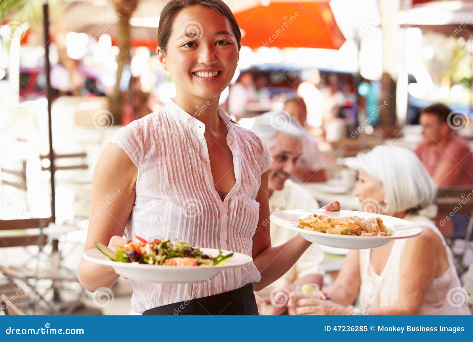  A smiling waitress wearing a white shirt and black apron carries two plates of food to customers sitting at a table outside a restaurant.