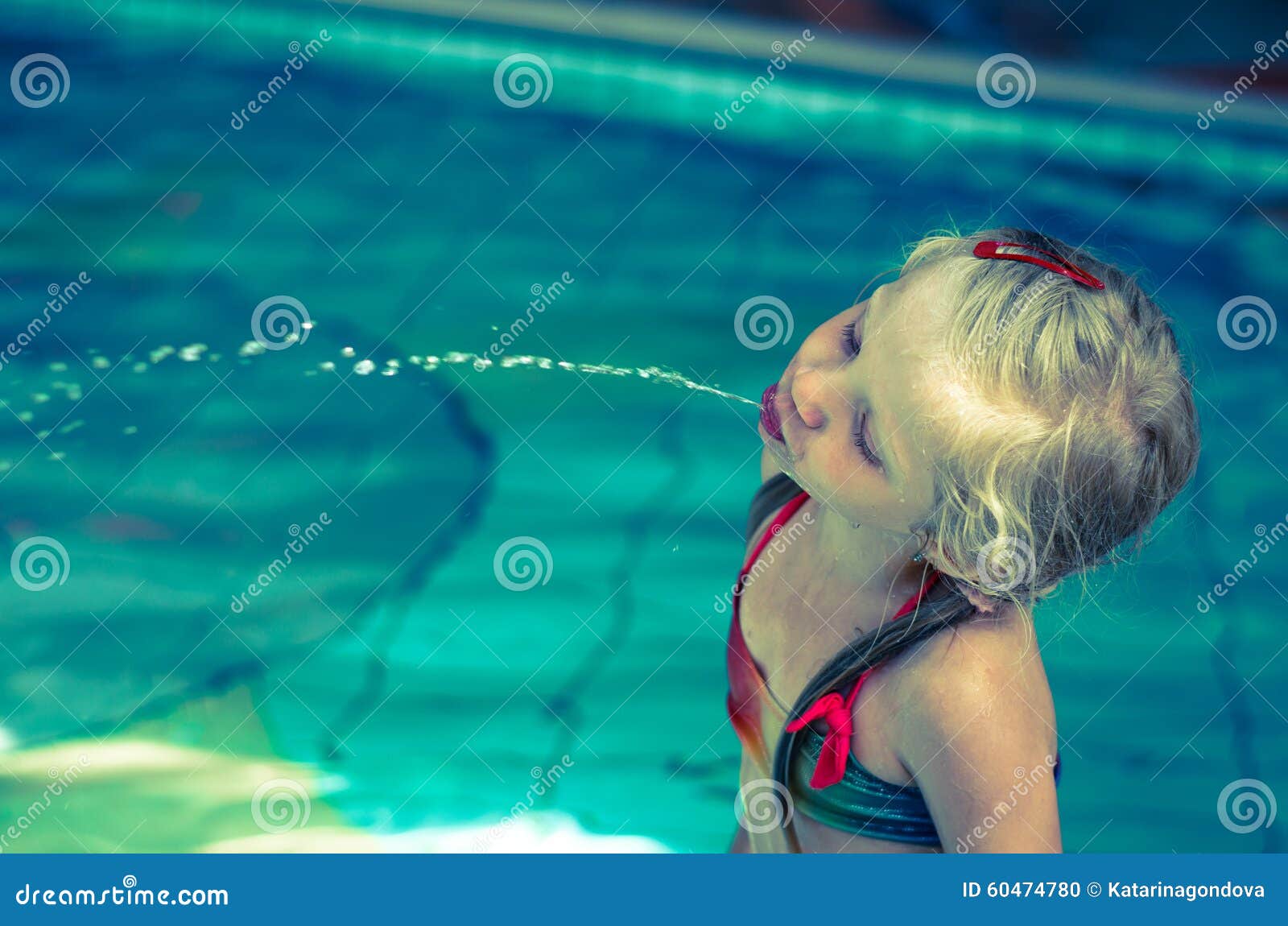 Water spray. Little blond girl in swimsuit spitting water in the swimming pool