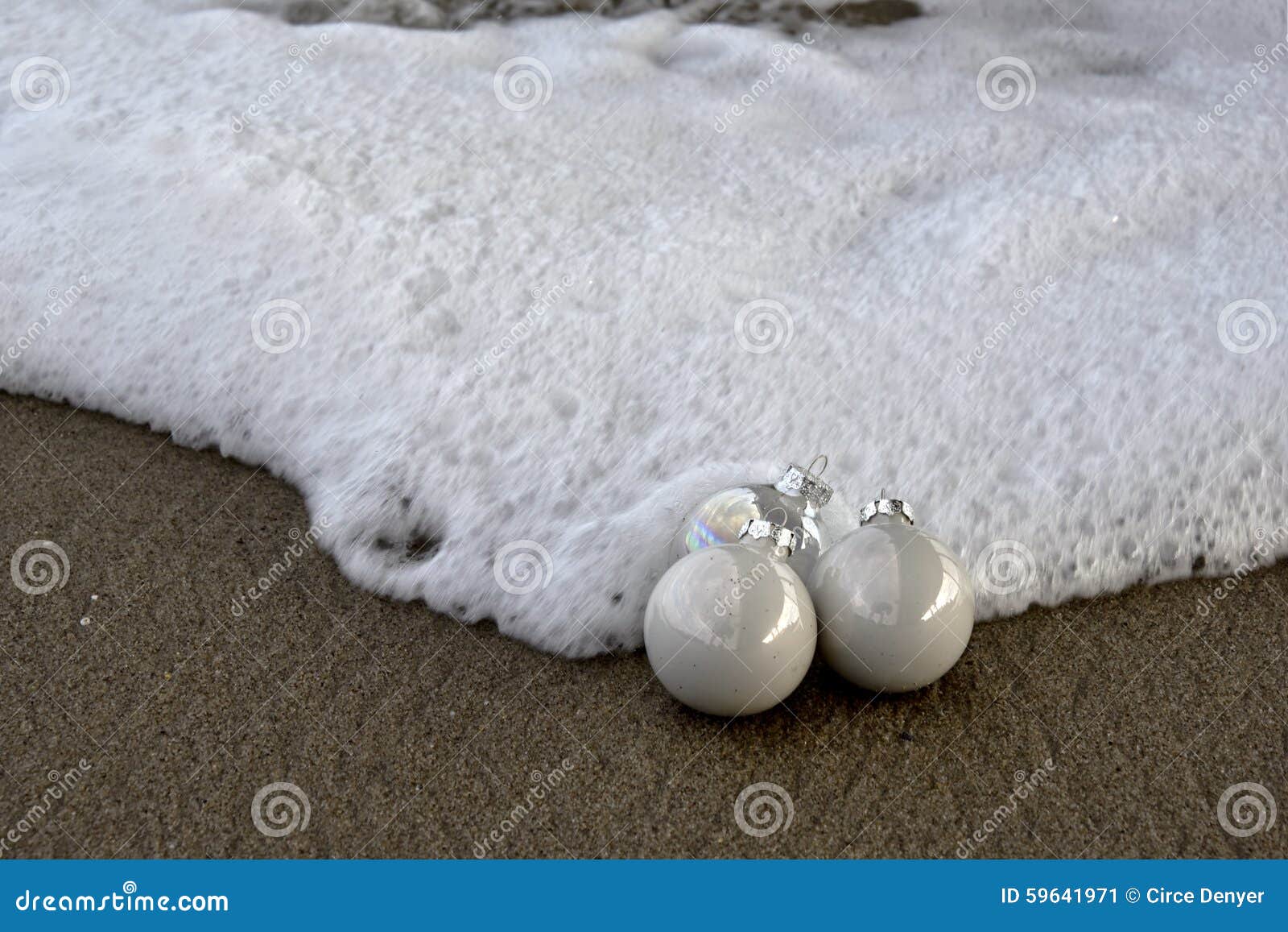 Weiche Gischt, die Weihnachtsverzierungen umarmt. Brandung und Sand umgeben diese drei Weihnachtsverzierungen auf dem Strand Weiße Verzierungen und weiße Brandung schaffen ein monotones Bild für Weihnachten