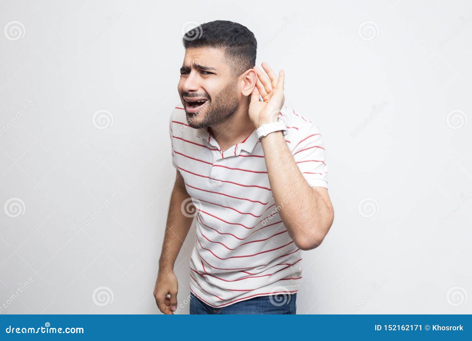 what? i can not hear you. portrait of attentive handsome bearded young man in striped t-shirt standing with hand on ear and want