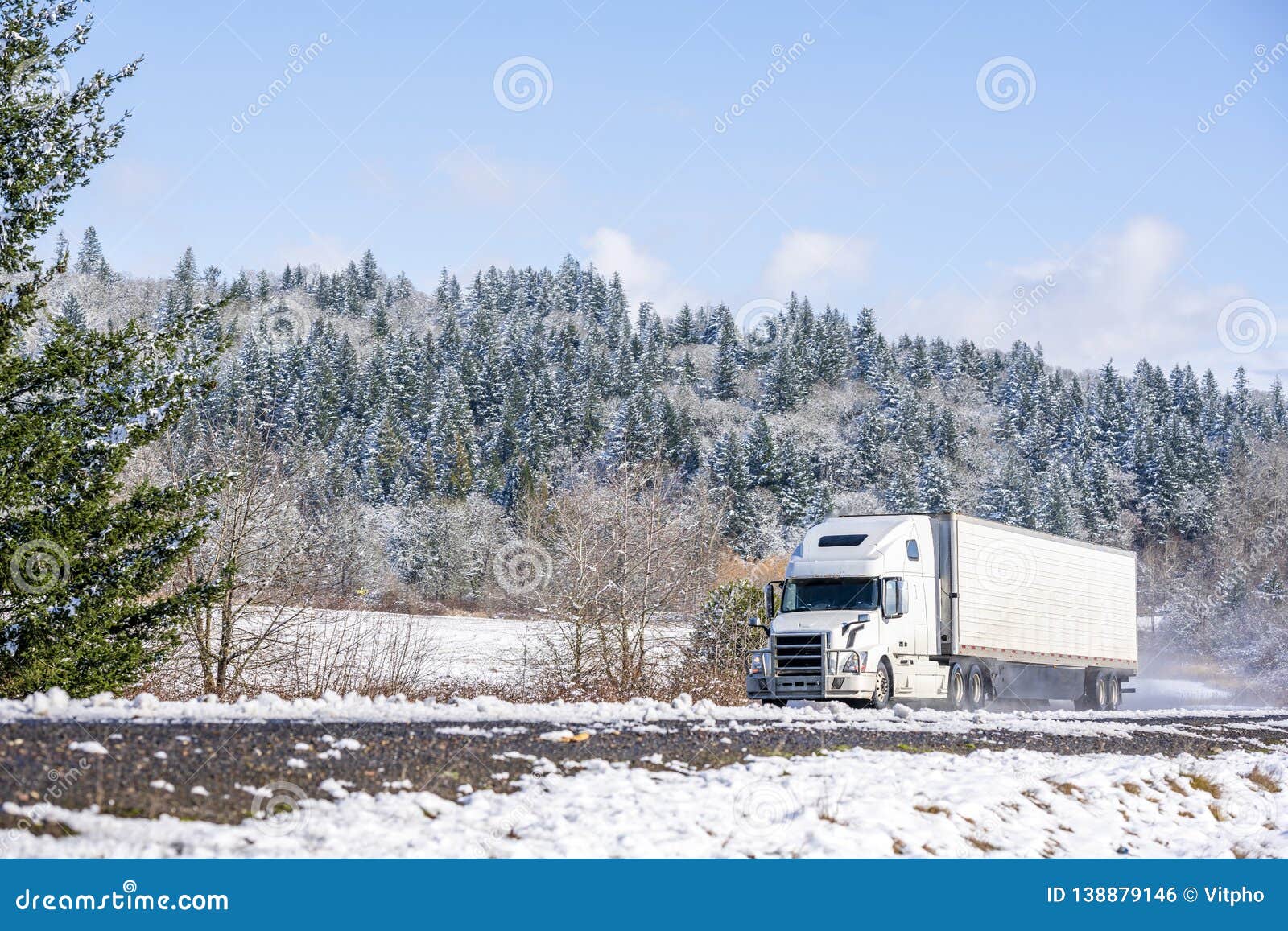 white big rig long haul semi truck with refrigerator semi trailer running on the winding winter snowy road with forest