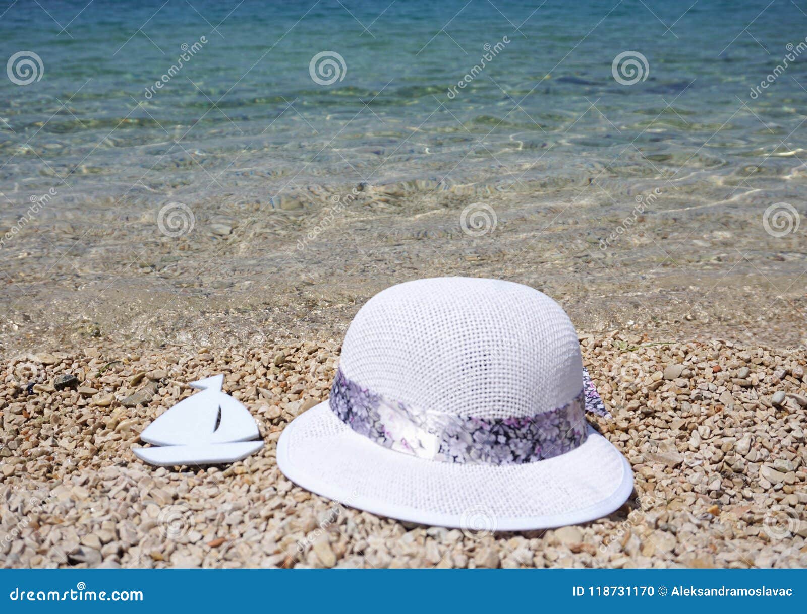 White boat from a paper and summer hat on the pebble beach with blue and turquoise sea in the background. White boat made from a paper and summer hat on the pebble beach with clear blue and turquoise sea in the background on a beautiful summer day.Summer vacation concept