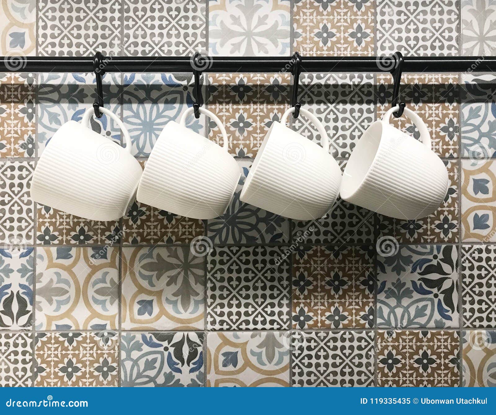 White Ceramic Cups Hanging on Hook in Front of Art Pattern Tile Stock