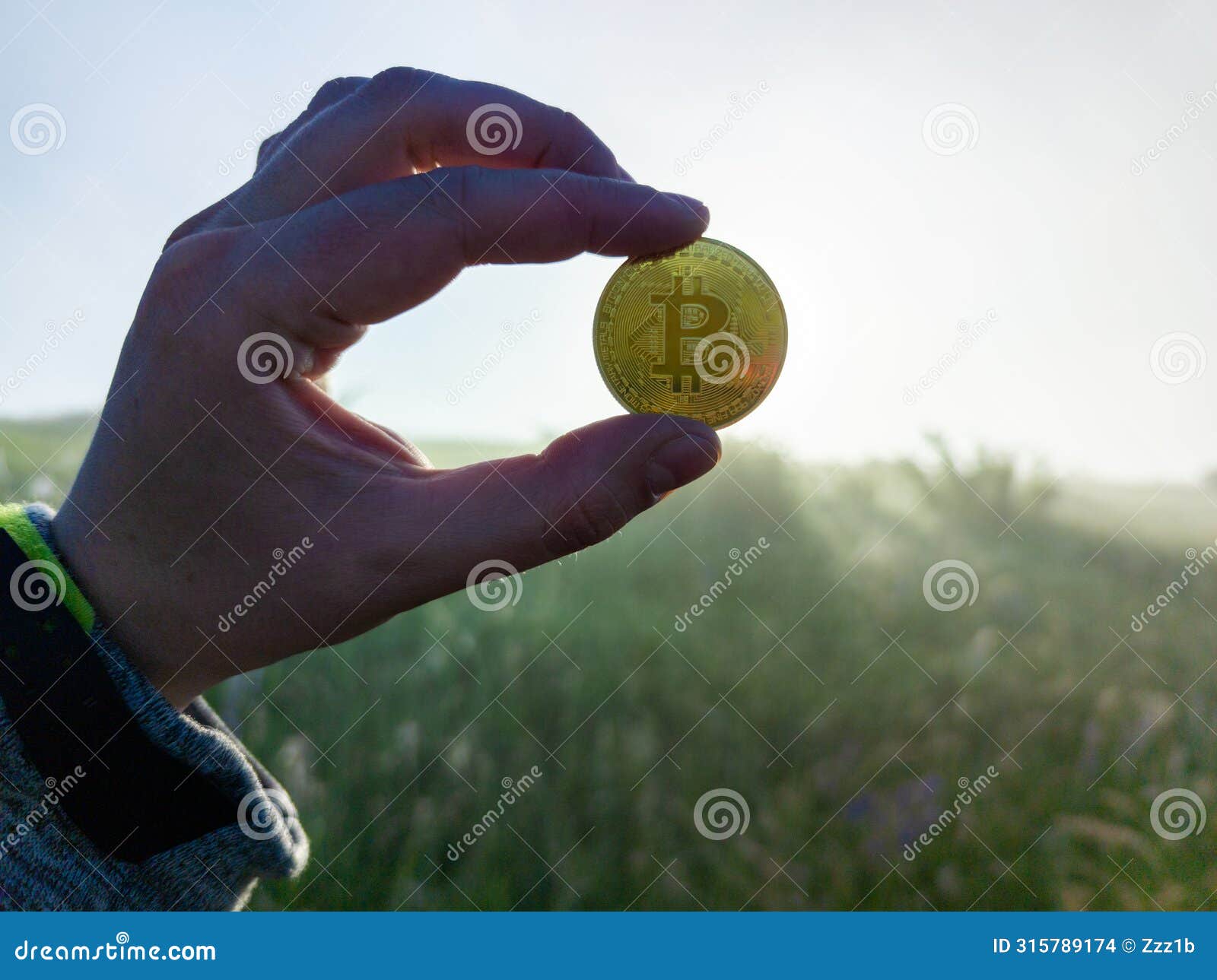 white human hand holding a bitcoin shiner on a morning grassy meadow background