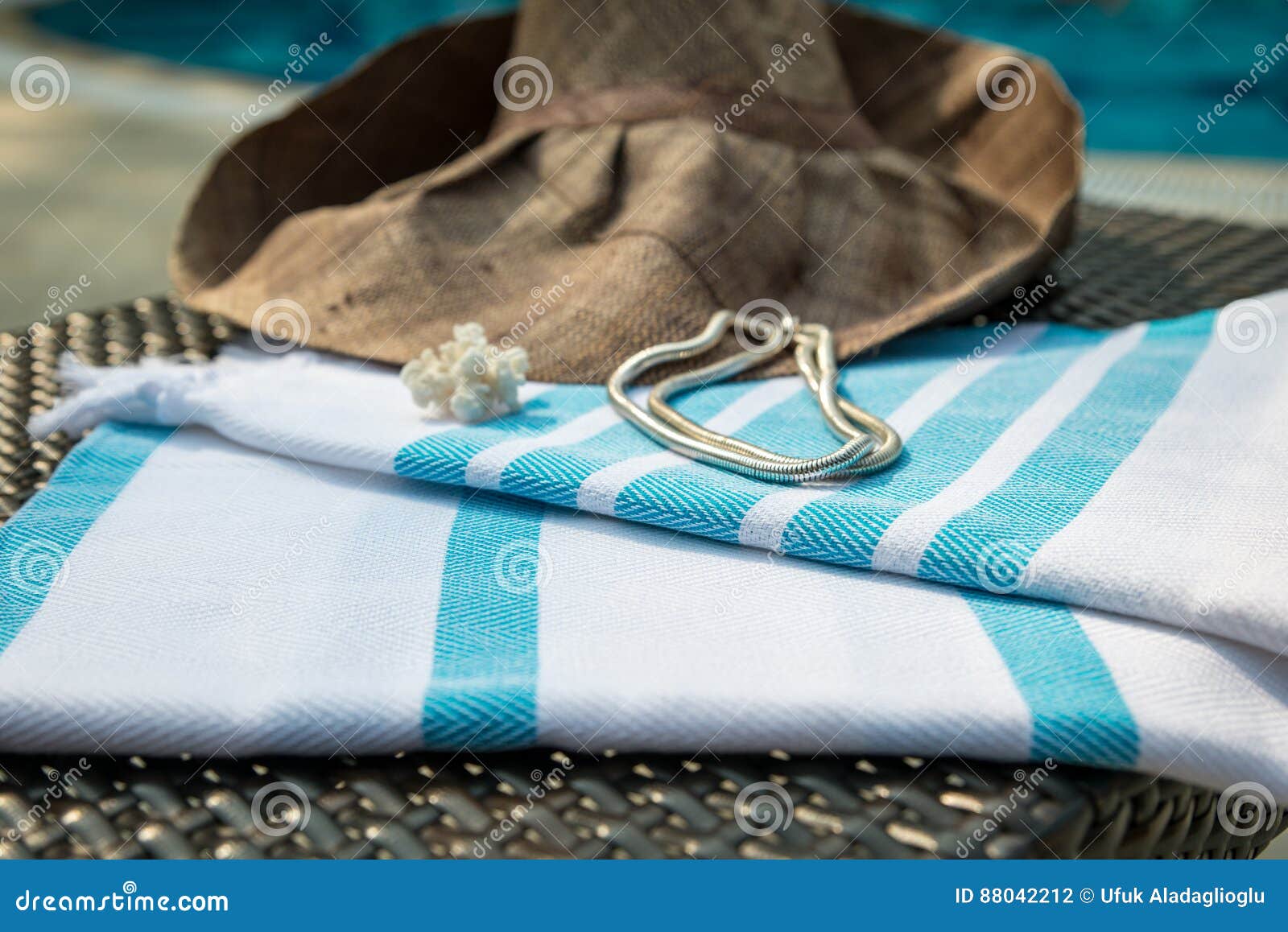 A white and turquoise color Turkish peshtemal, white seashells, white gold necklace and straw hat on rattan lounger. The concept of summer accessories close-up of white and turquoise color Turkish peshtemal, white seashells, white gold necklace and straw hat on rattan lounger with a blue swimming pool as background