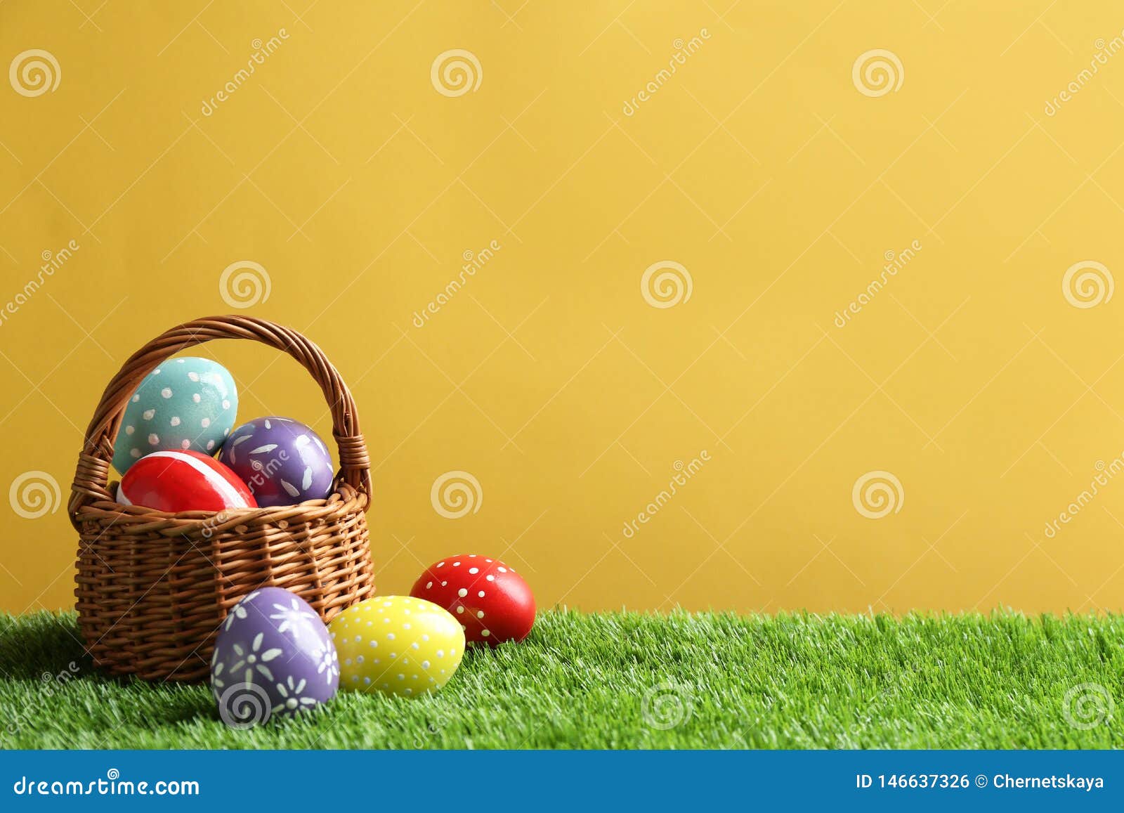 Wicker Basket with Painted Easter Eggs on Green Grass Against Color