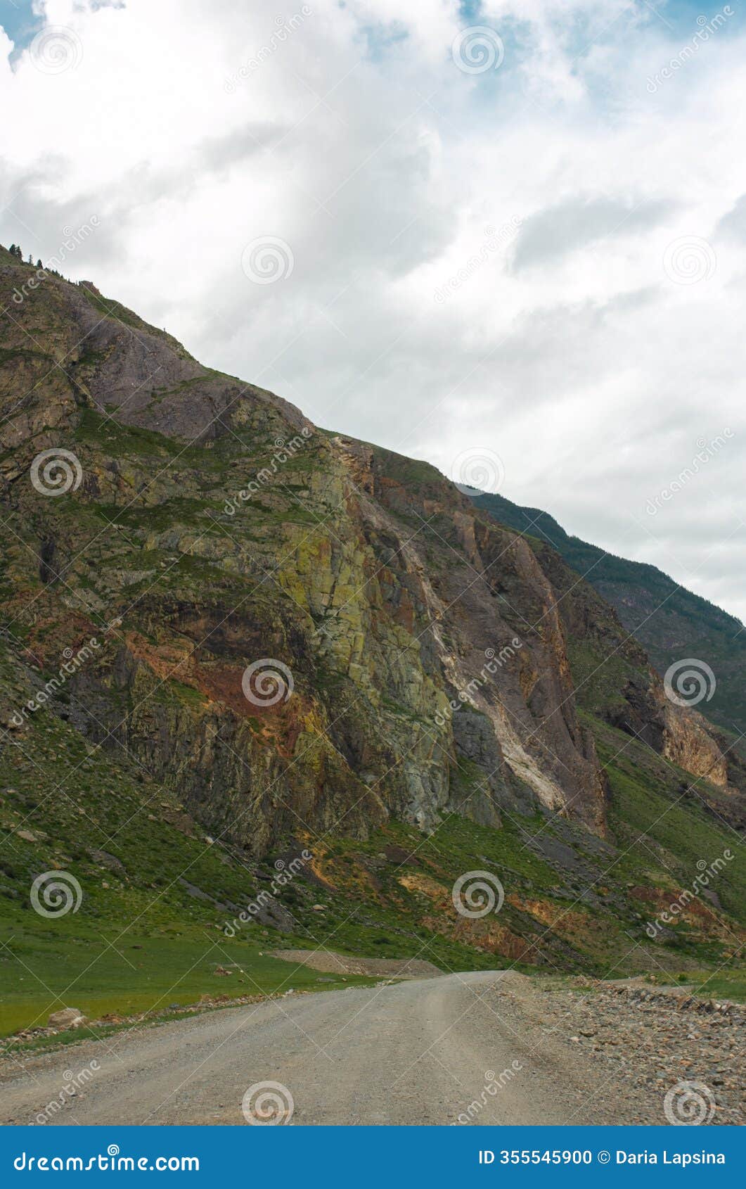winding gravel road cuts through the rugged landscape of mountain valley under cloudy sky. towering cliffs and lush