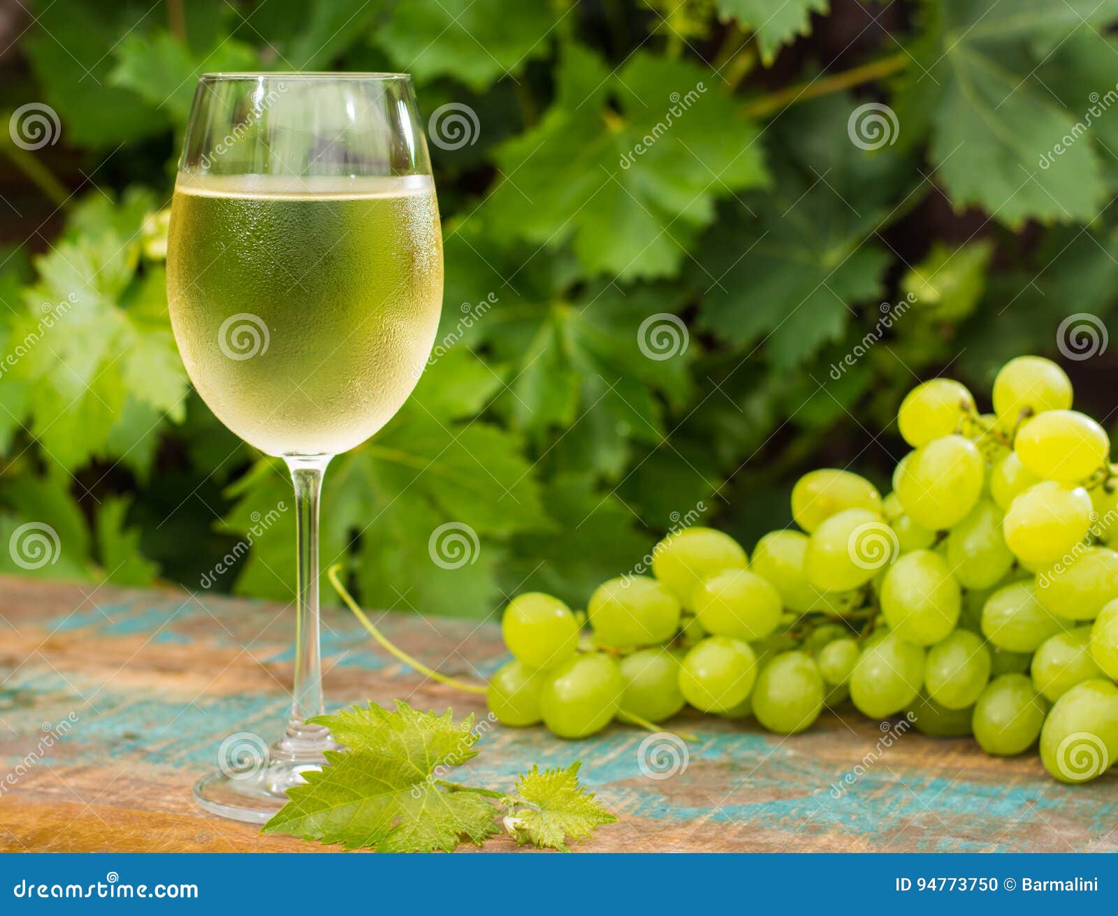 Wine glass with ice cold white wine, outdoor terrace, wine tasting in sunny day, green vineyard garden background and white grape