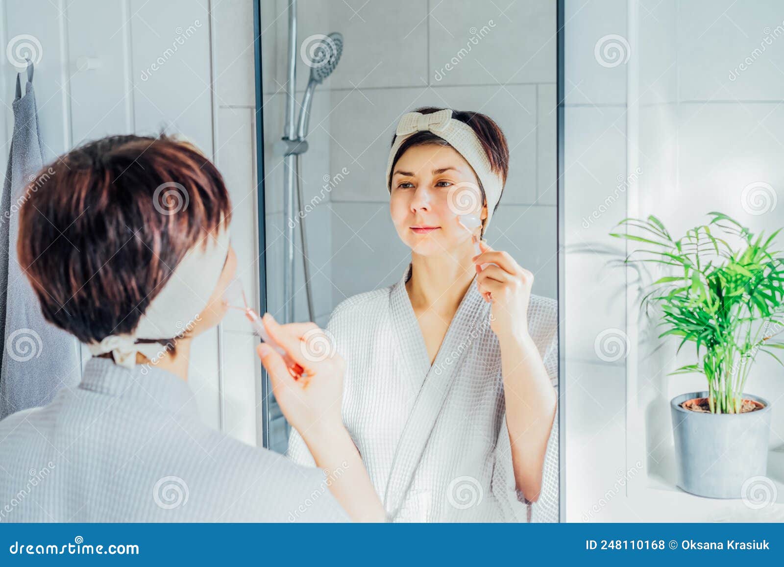 Woman In Bathrobe And Hair Band Looking In The Mirror And Making Face Massage With Jade Roller