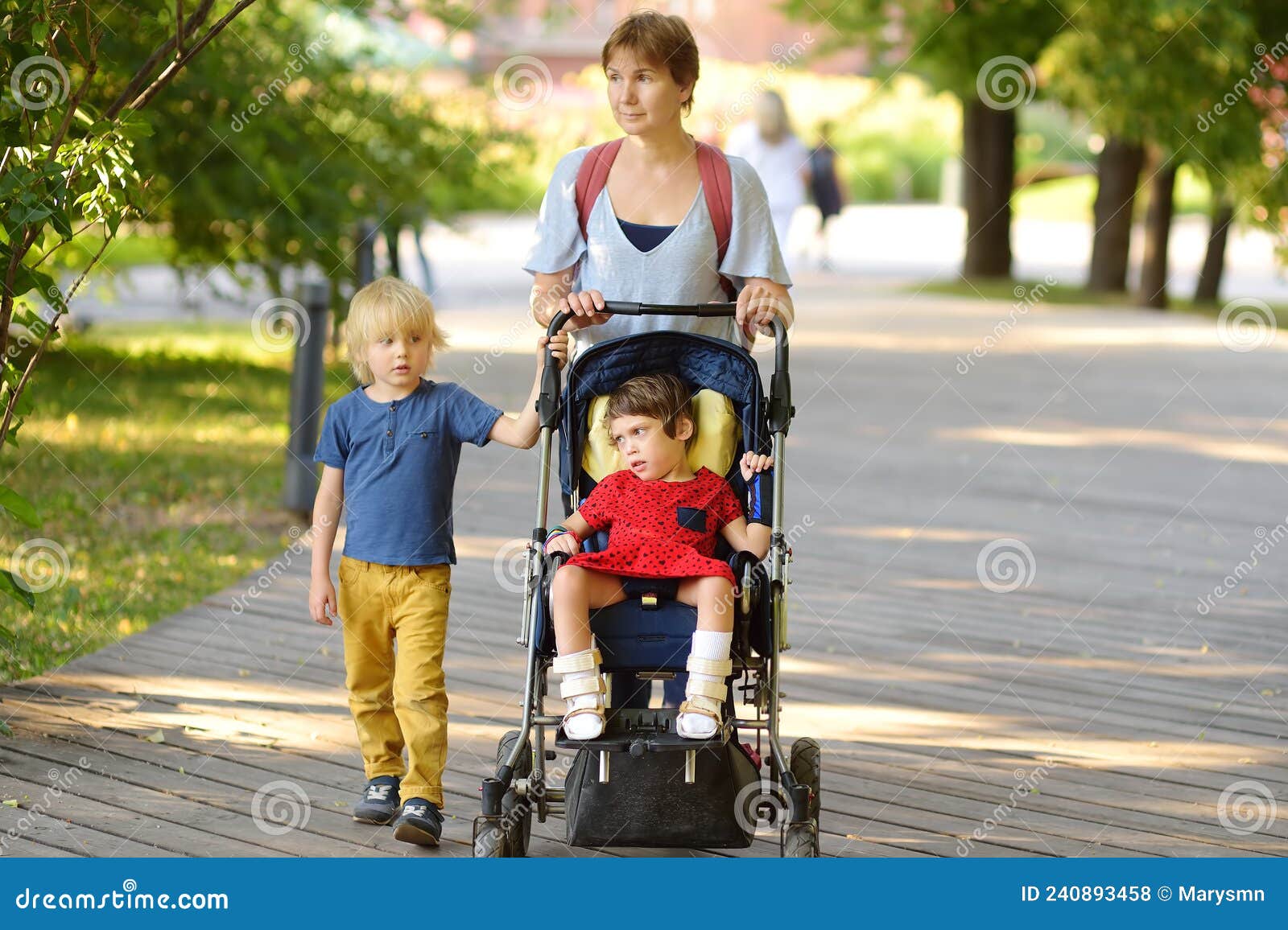 woman with a boy and a disabled girl in wheelchair walking in park summer. on baby legs orthosis. child cerebral palsy. treatment