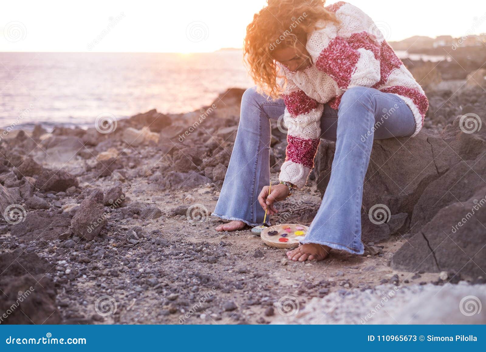 woman with curly hair and flared trousers and rock rodipinge sweater at sunset sitting on a rock at an  beach