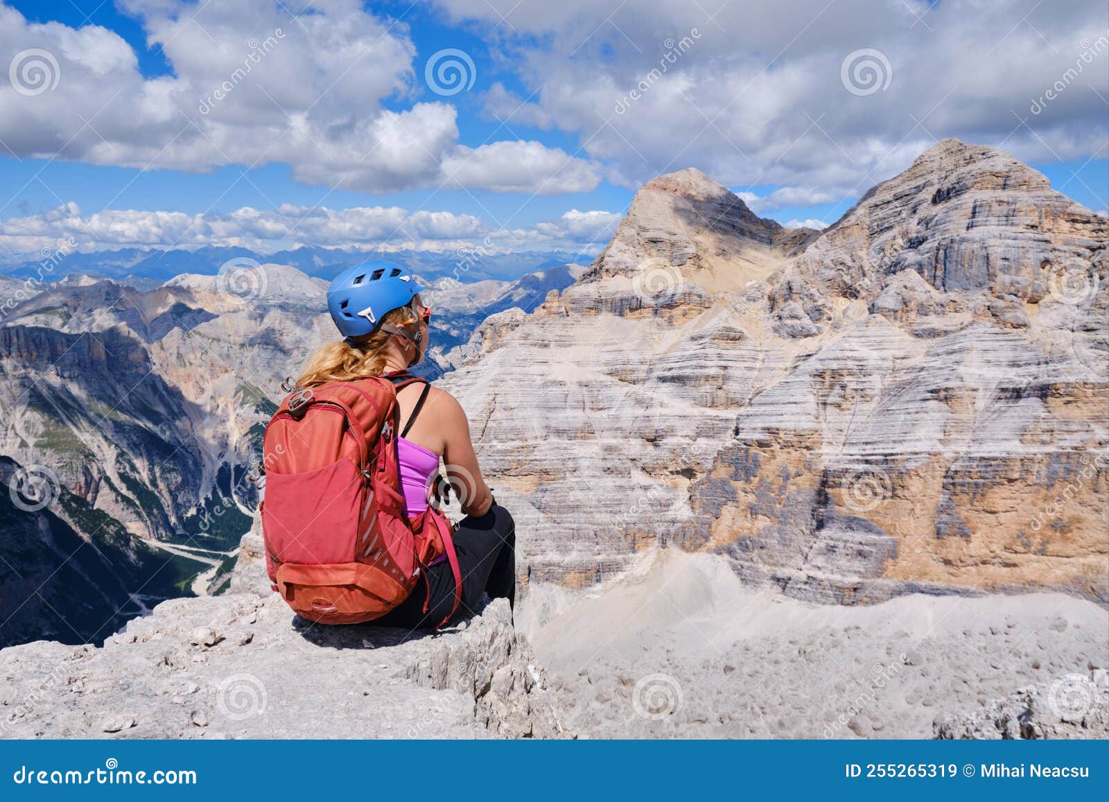woman hiker with helmet and red backpack looks towards tofana di mezzo and di dentro in dolomites mountains, italy.