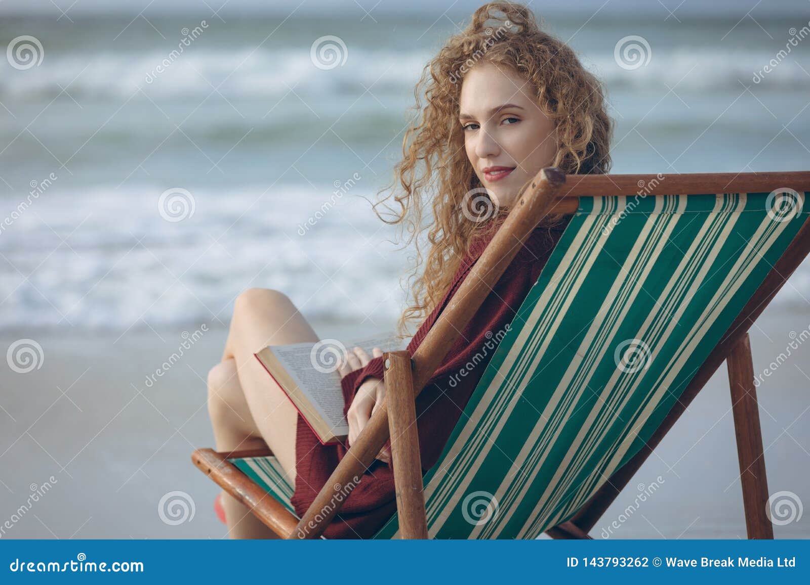 Woman Holding Book While Sitting On Sun Lounger At Beach Stock Photo