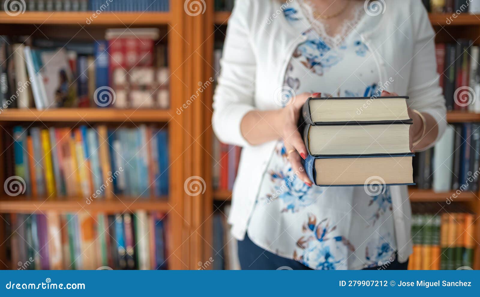 Woman Holding a Stack of Large Books with a Large Bookcase Full of