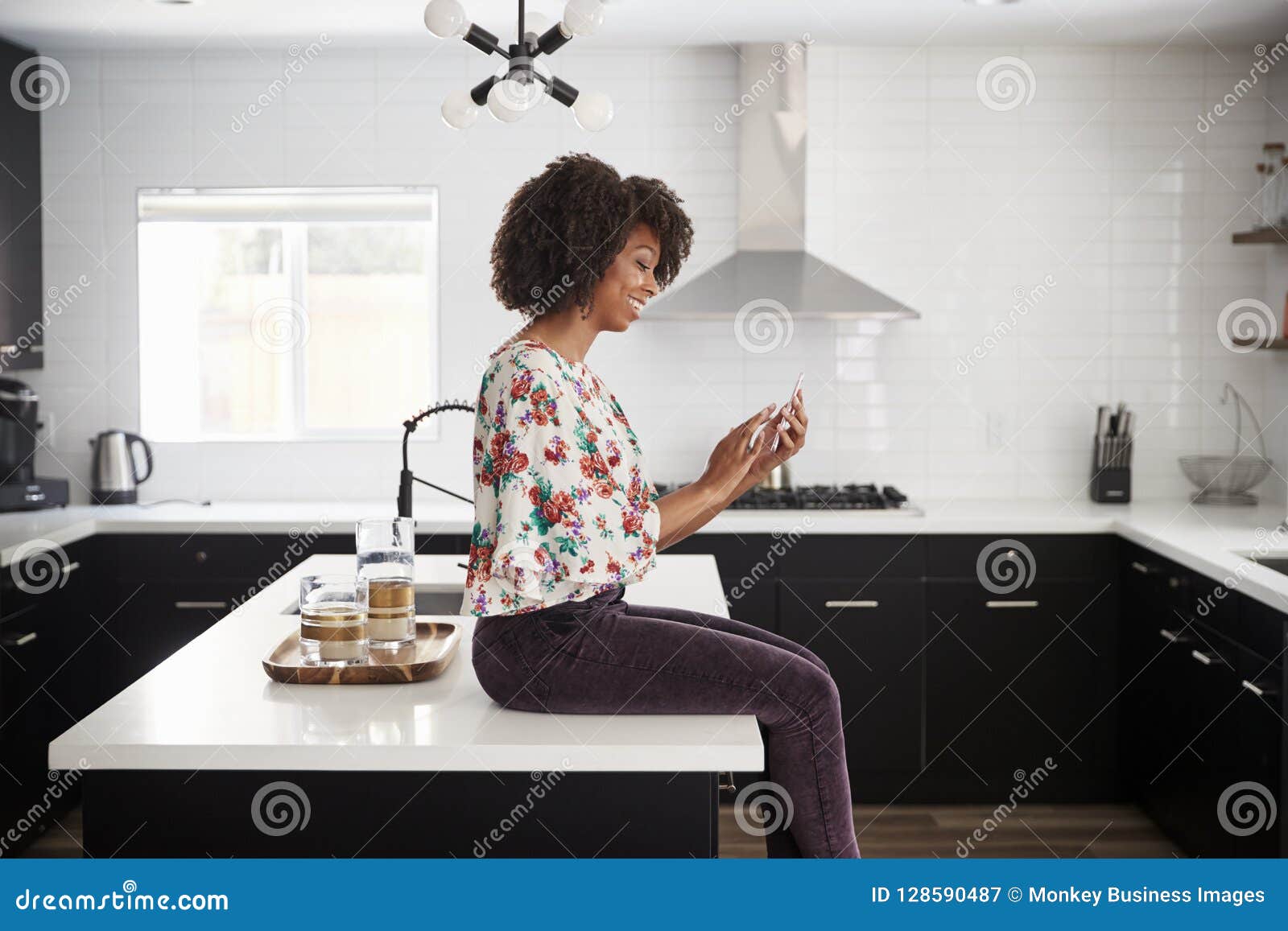 Woman At Home Sitting On Kitchen Island Whilst Using Mobile Phone