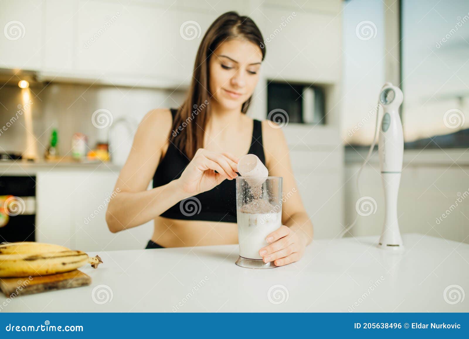 woman with immersion blender making banana chocolate protein powder milkshake smoothie.adding a scoop of low carb whey protein mix