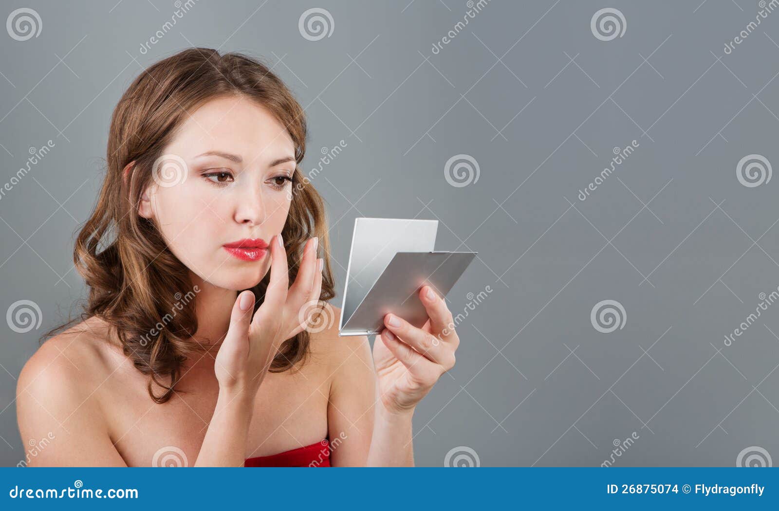 Woman with mirror. Beautiful young woman having make-up and looking in the mirror. Studio shot. Empty space for text. Gray background.