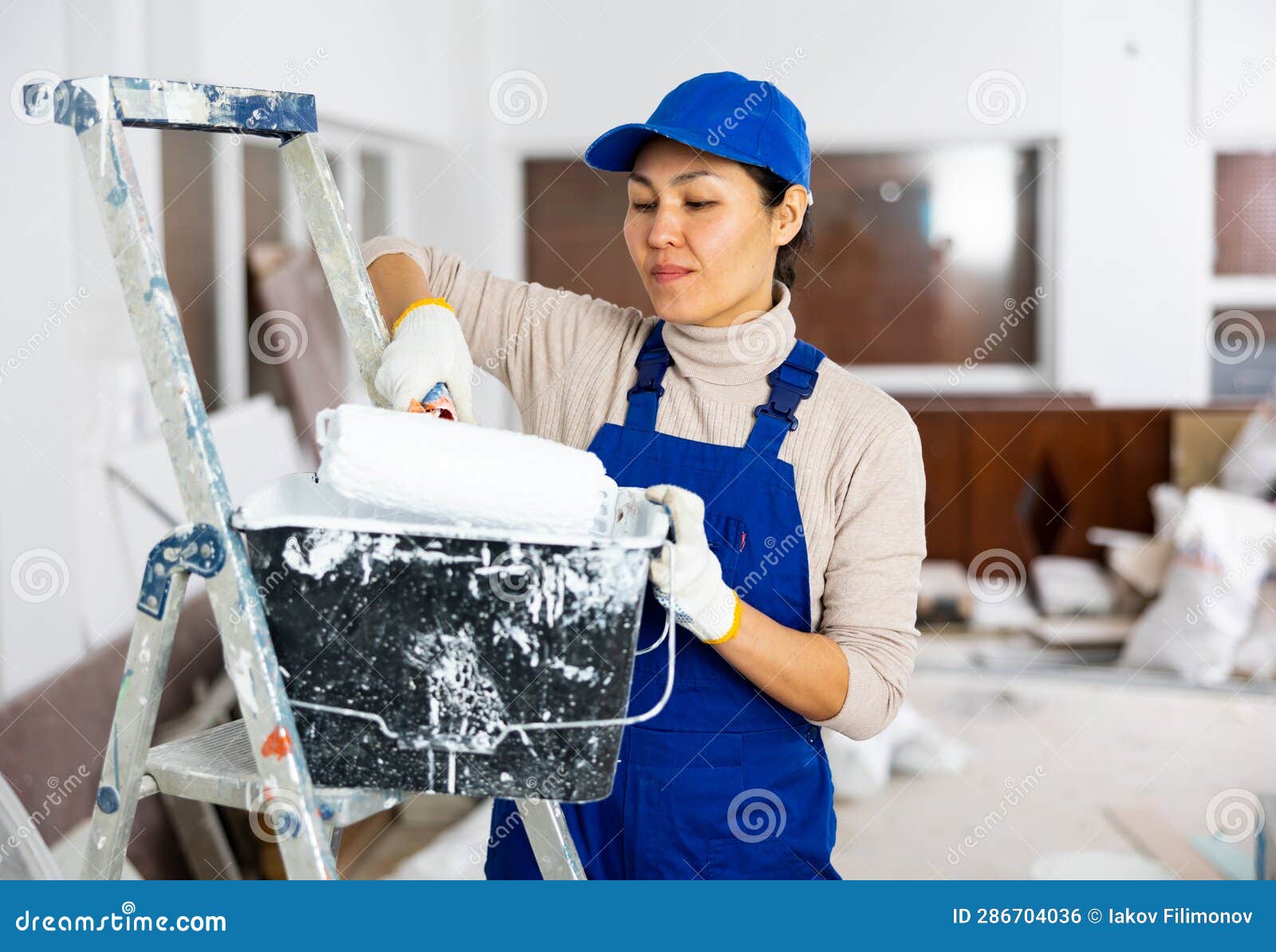 Woman Painter in Overalls with Paint Roller in Building Under