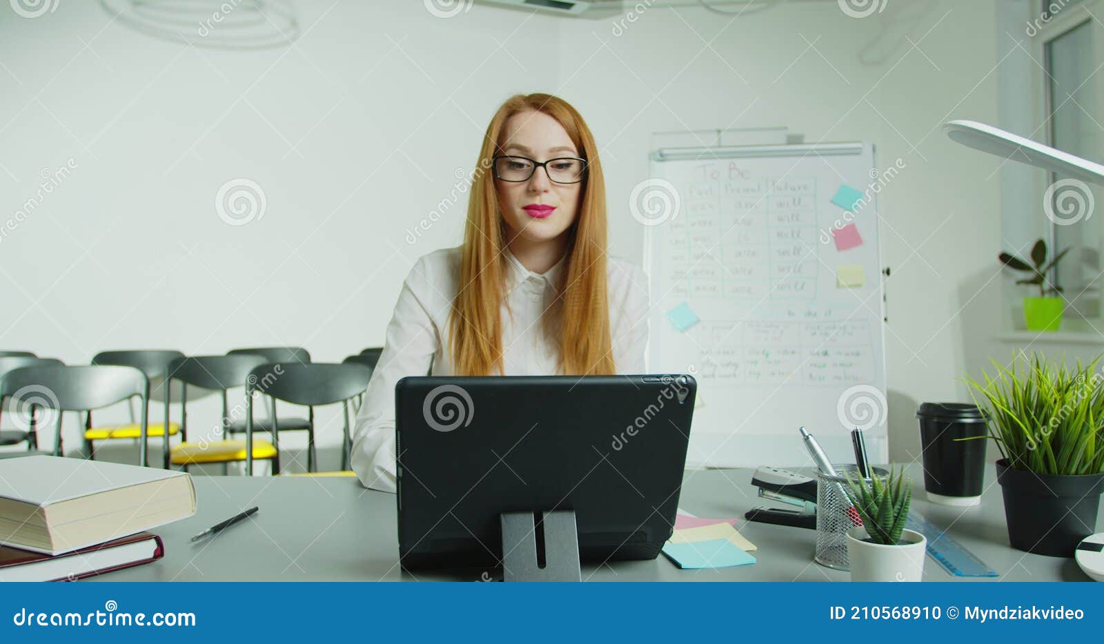 woman professor sitting at table and lecturing online at class. female english teacher in glasses speaking on tabletpc