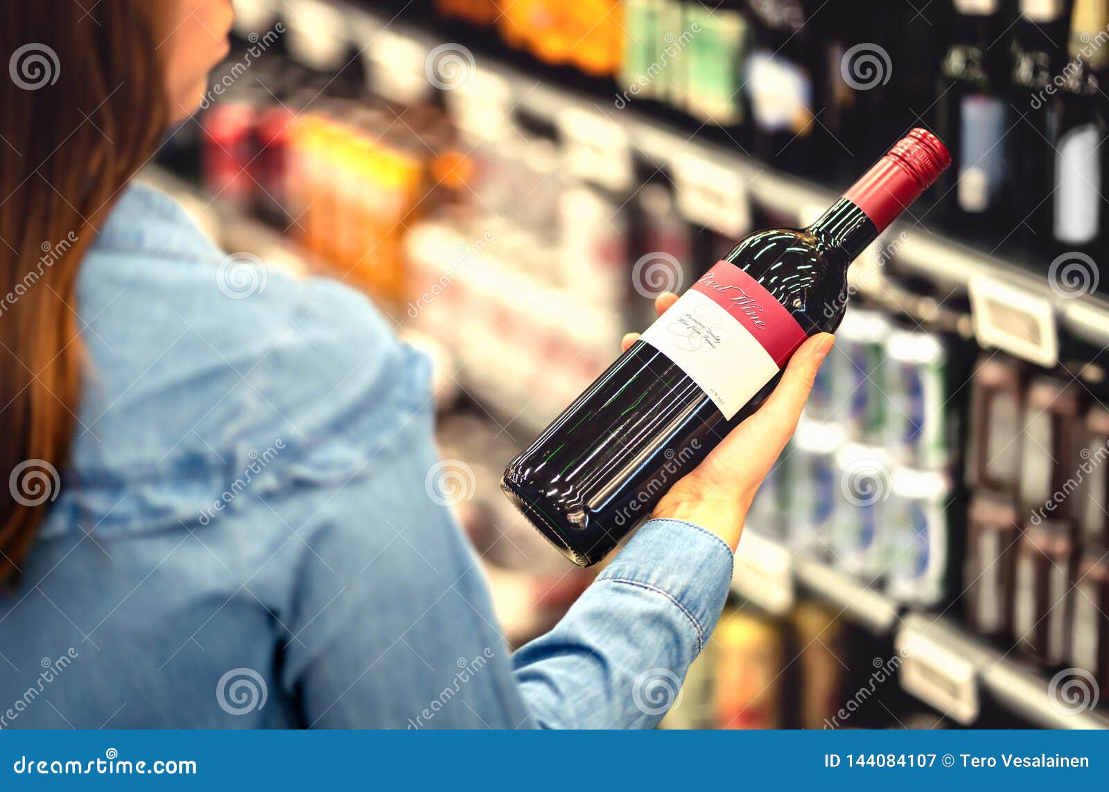woman reading the label of red wine bottle in liquor store or alcohol section of supermarket. shelf full of alcoholic beverages.