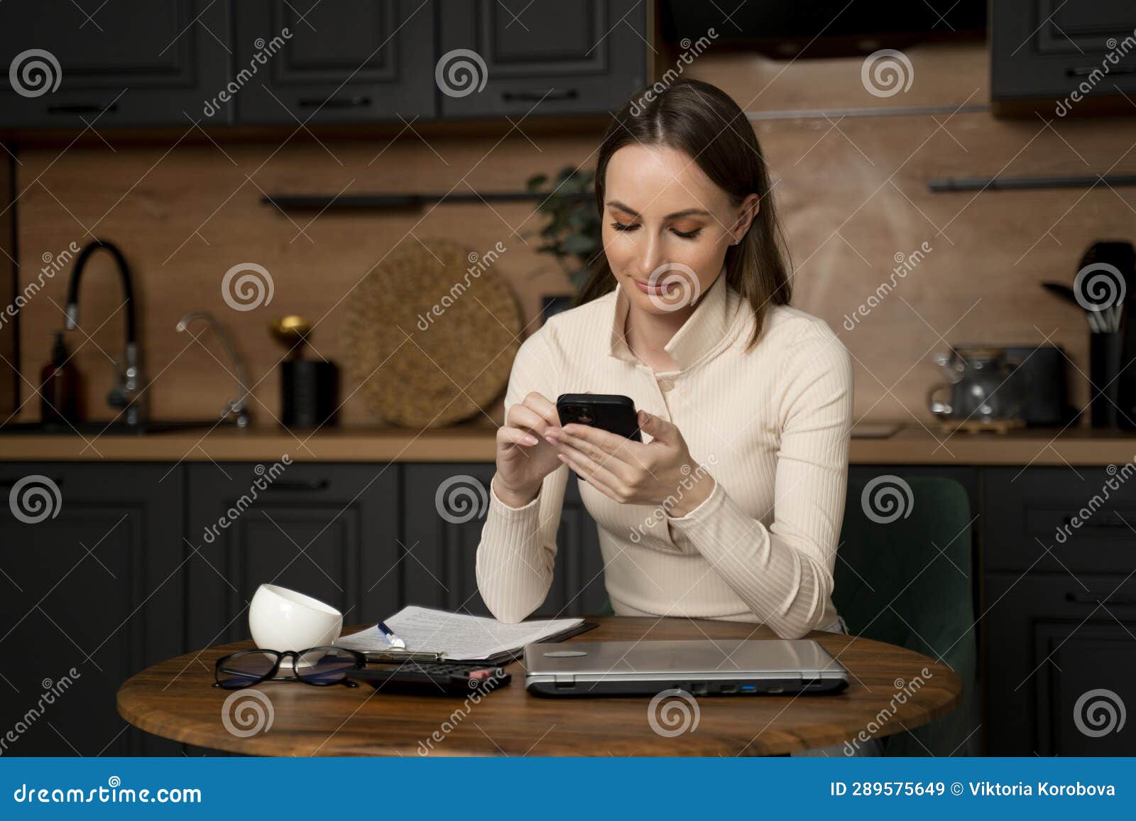 Woman Sitting at the Kitchen Table with a Mobile Phone is Using and