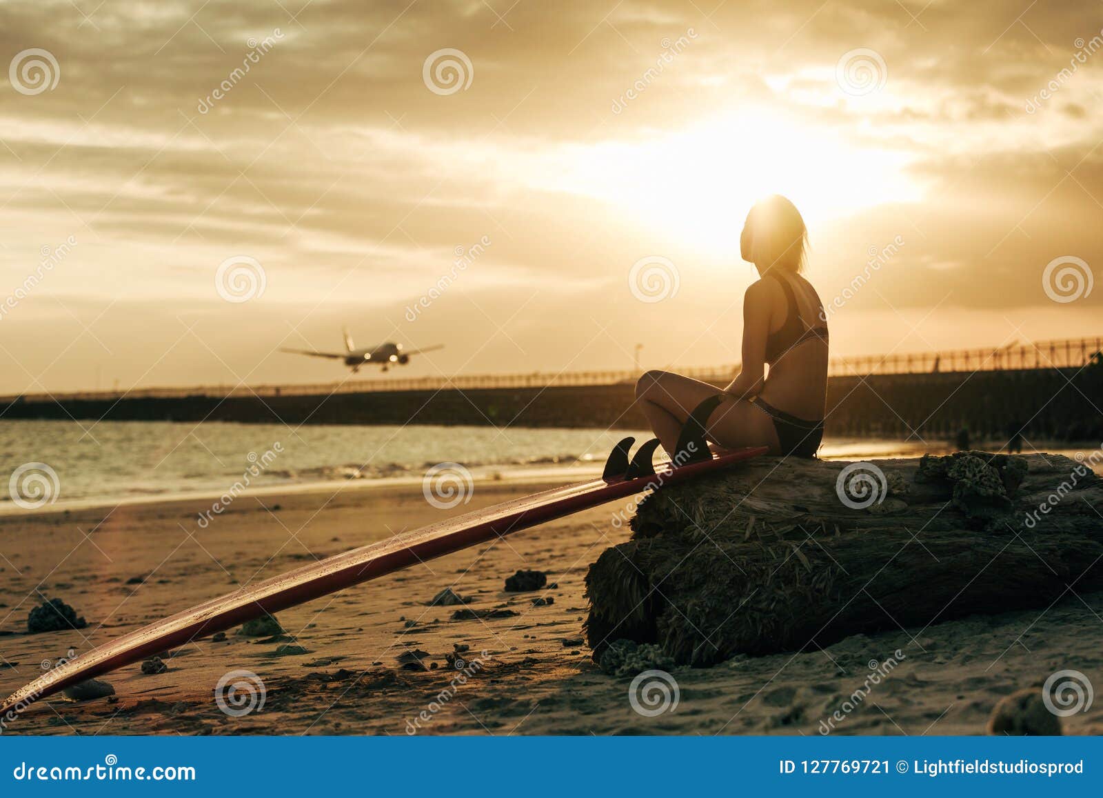 Woman Sitting On Rock With Surfboard On Beach At Sunset With Airplane