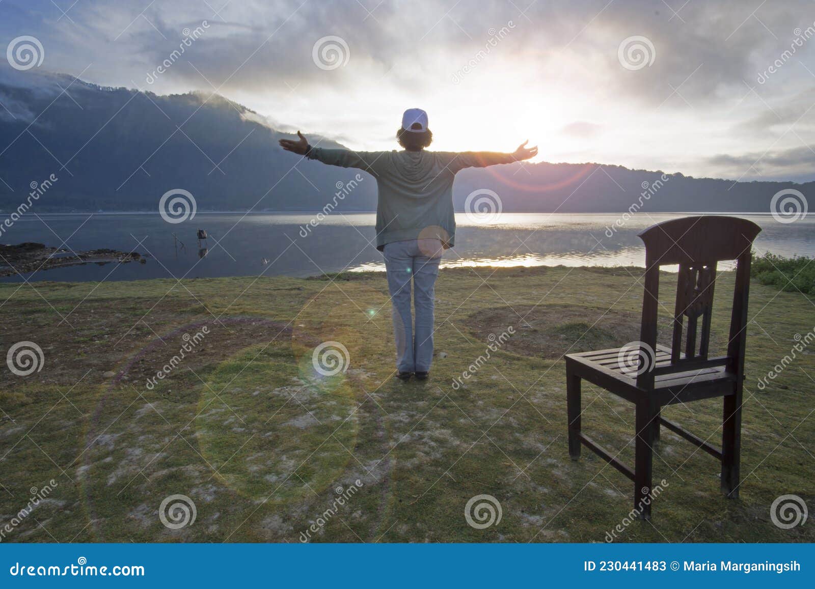 woman standing alone on the ground, hands raise with open arms against the light over the lake.