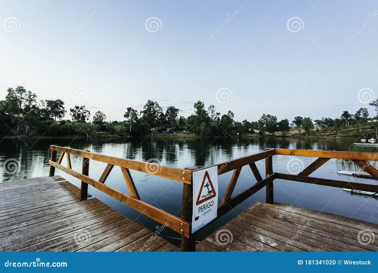 wooden dock with a [perigo - danger] sign on a lake under a blue sky and sunlight at daytime