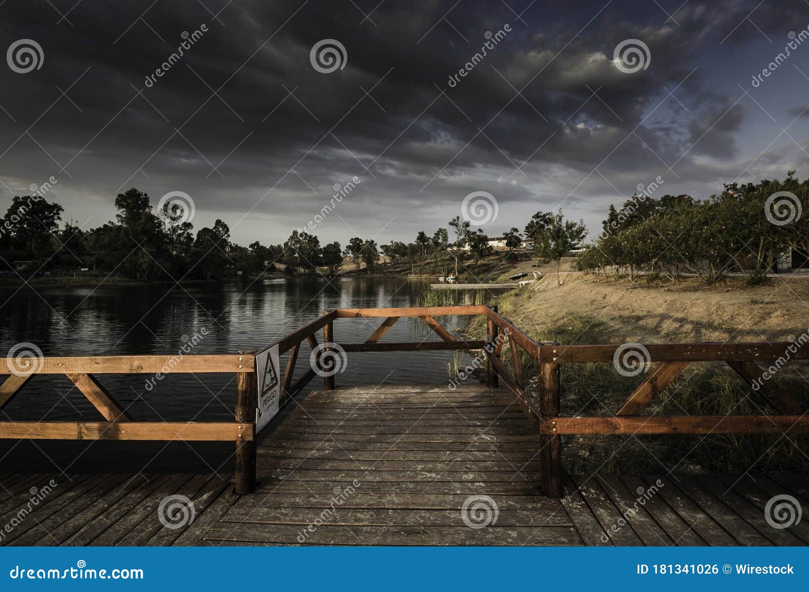 wooden dock with a [perigo - danger] sign on a lake under a dark cloudy sky in the evening