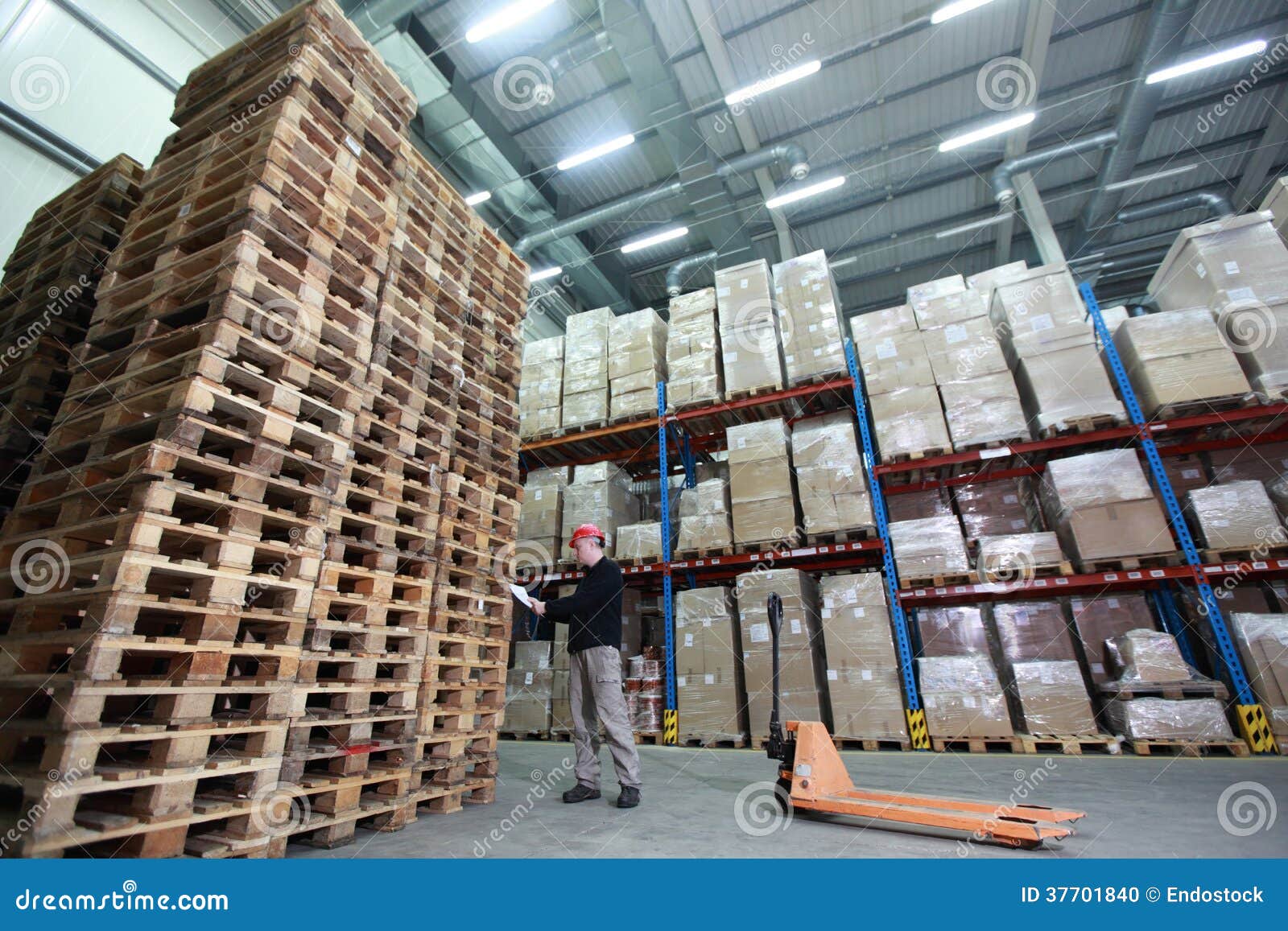 worker with hand pallet truck at large stack of wooden pallets in storehouse