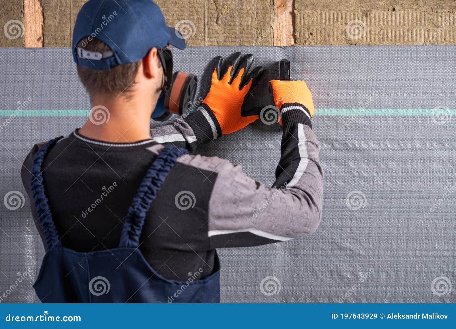 a worker in overalls, using a stapler, mounts a vapor barrier membrane to protect the mineral insulation
